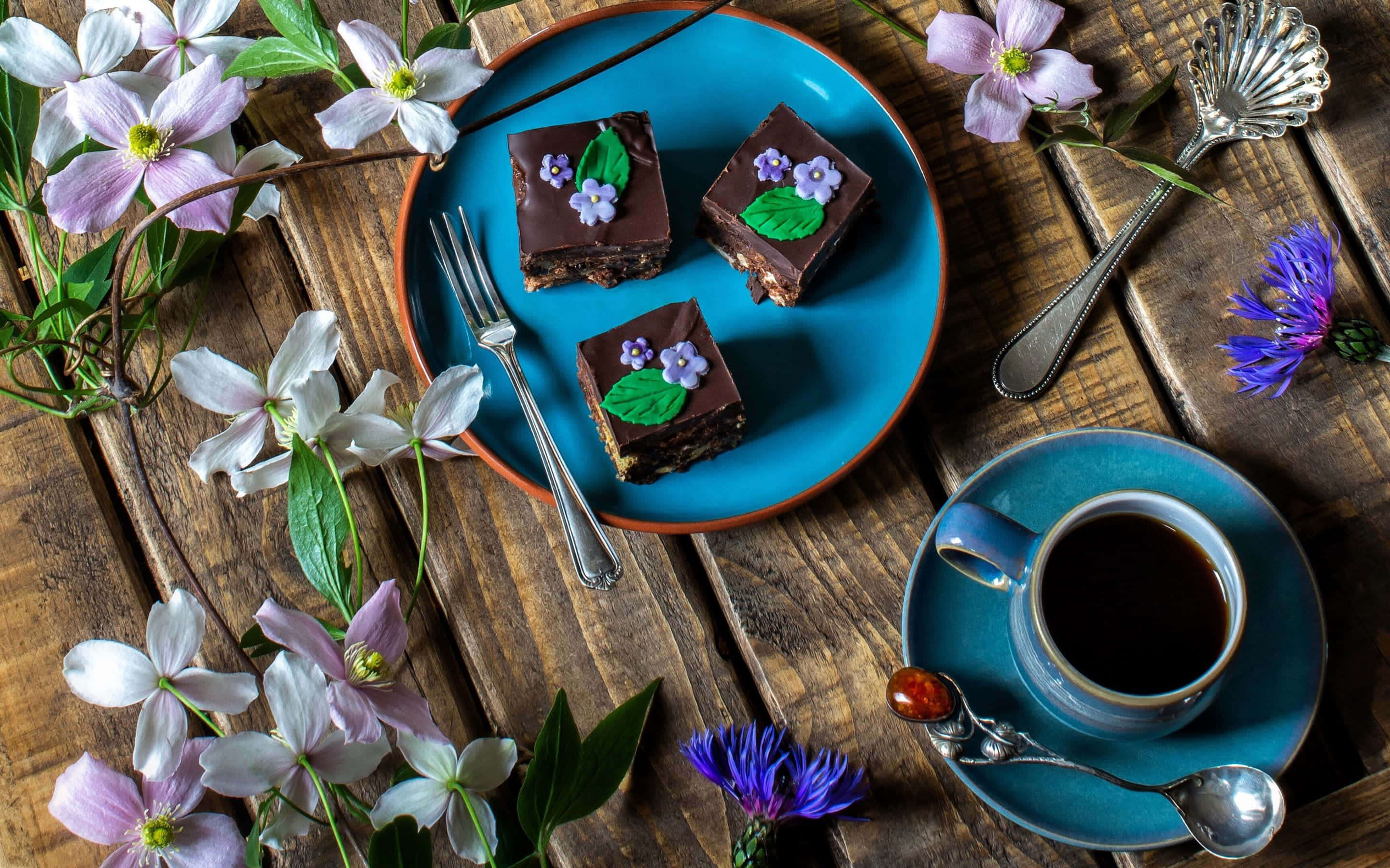 Spring Desserts 桌布 壁紙 — 三塊飾有食用花的巧克力蛋糕方塊放在藍色盤子上，旁邊有一杯咖啡，置於鄉村風格的木桌上。