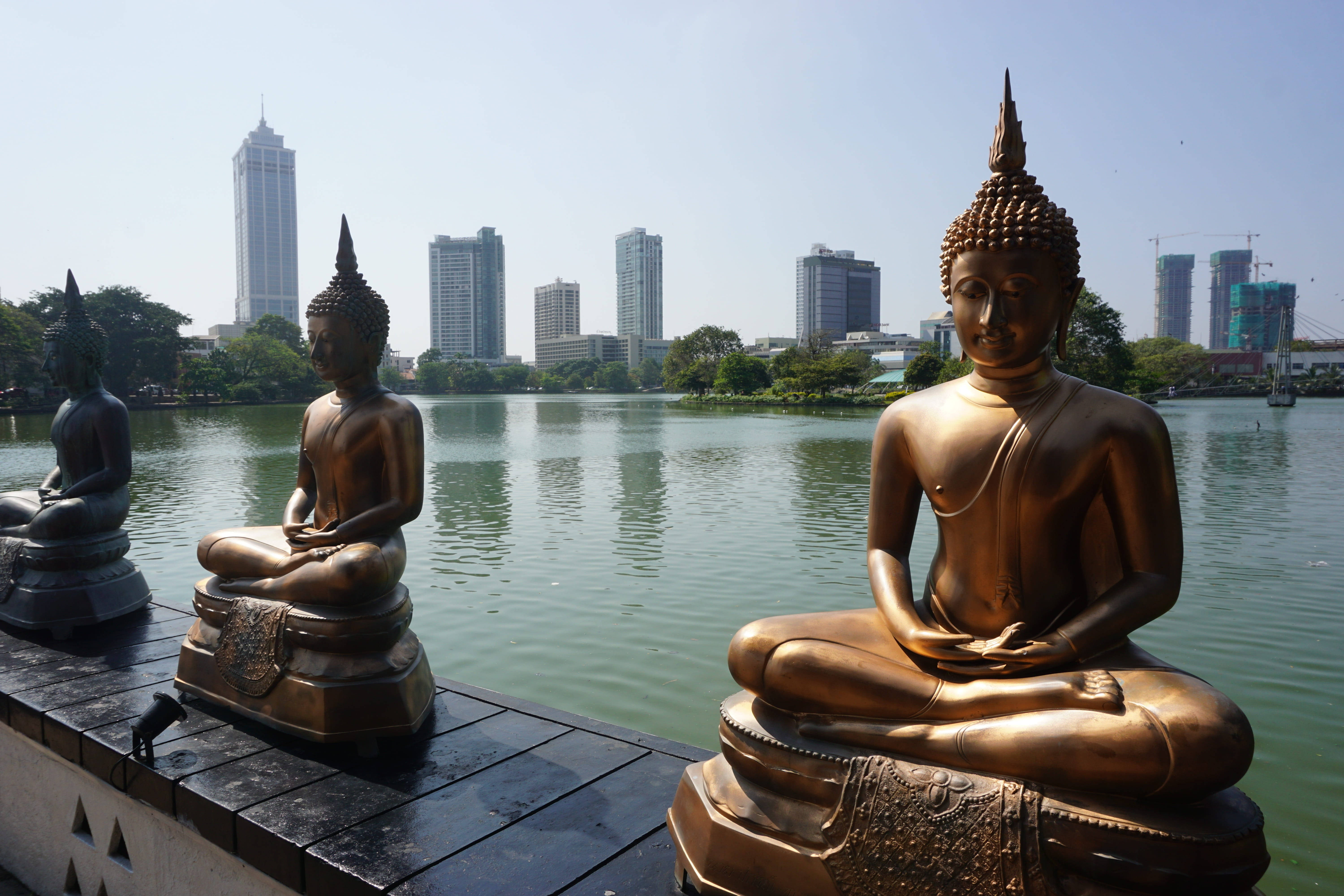 Sri Lanka 桌布 壁紙 — 三尊寧靜的佛像在湖邊碼頭上打坐，現代摩天大樓在晴朗的天空下倒映在平靜的水面上。