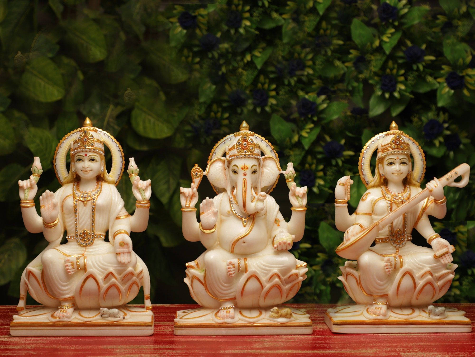 Hindu God 桌布 壁紙 — 三尊精雕細琢的白色大理石印度教神像，寧靜地坐在紅色木質表面上，背景是茂密的綠色樹葉。
