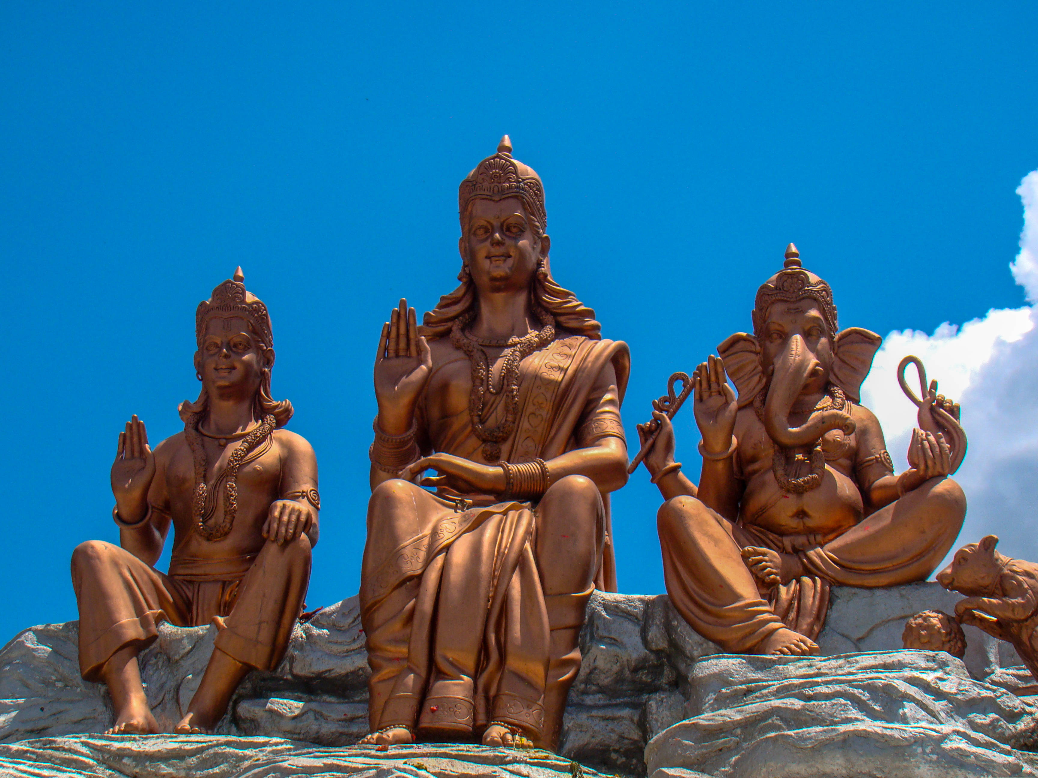 Bappa 桌布 壁紙 — 三尊青銅印度教神像在岩石地形上寧靜地坐著，背景是鮮豔的藍天和蓬鬆的白雲。