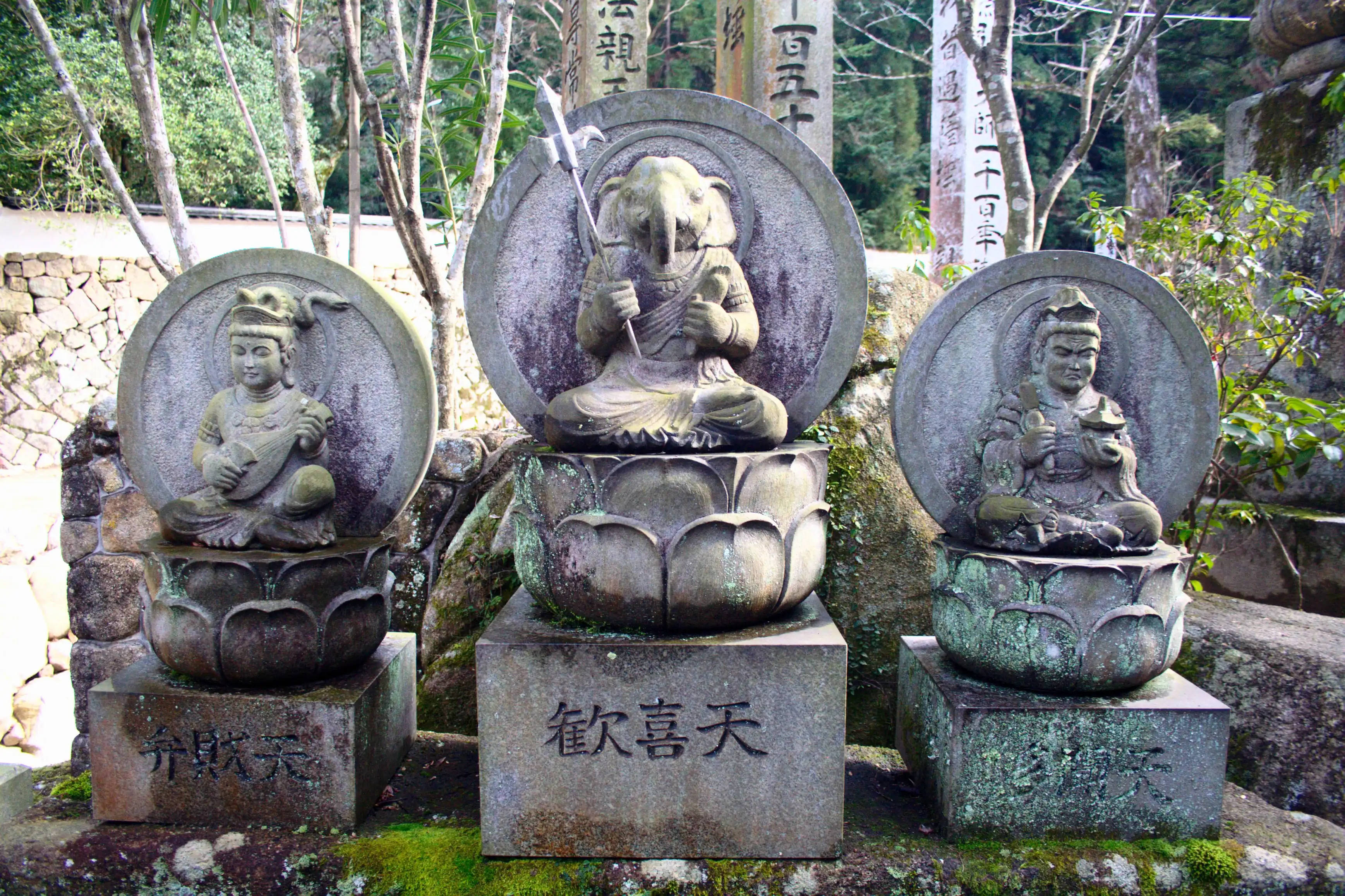 毗濕奴 桌布 壁紙 — 三尊風化的佛教神靈石像，安詳地坐在蓮花座上，周圍是長滿青苔的岩石和樹木，位於寧靜的寺院花園中。