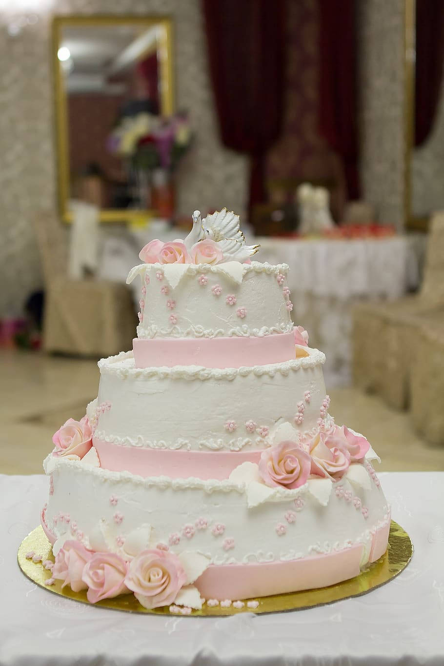 Wedding 桌布 壁紙 — 三層婚禮蛋糕，飾有粉紅玫瑰和白色糖花，優雅地放在宴會廳的金色底座上。