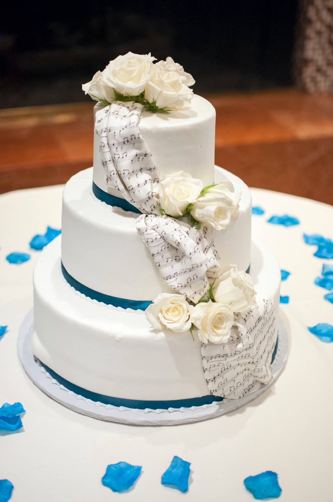 Wedding 桌布 壁紙 — 三層白色結婚蛋糕，裝飾著白玫瑰和樂譜絲帶，周圍散落著藍色花瓣在白色桌子上。