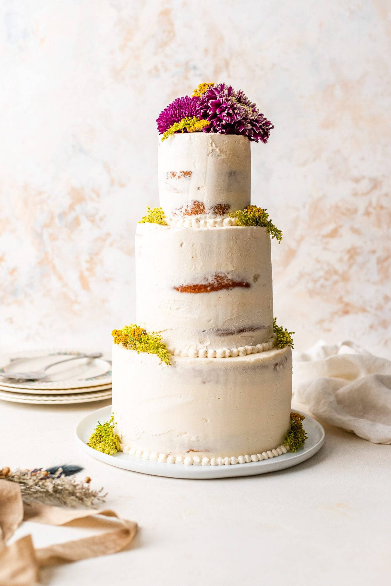 Wedding 桌布 壁紙 — 三層白色蛋糕，帶有鄉村風格的糖霜和鮮豔的紫色花朵，優雅地放在盤子上，旁邊有亞麻布和盤子。