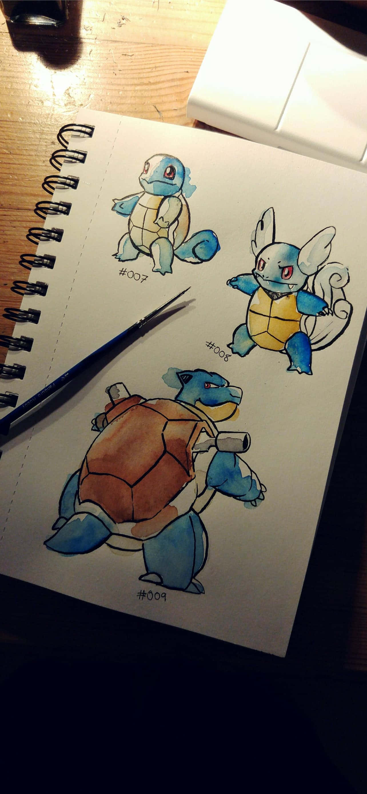 Squirtle 桌布 壁紙 — 三幅水彩寶可夢草圖畫在螺旋筆記本上，附近有一支畫筆放在木桌上。