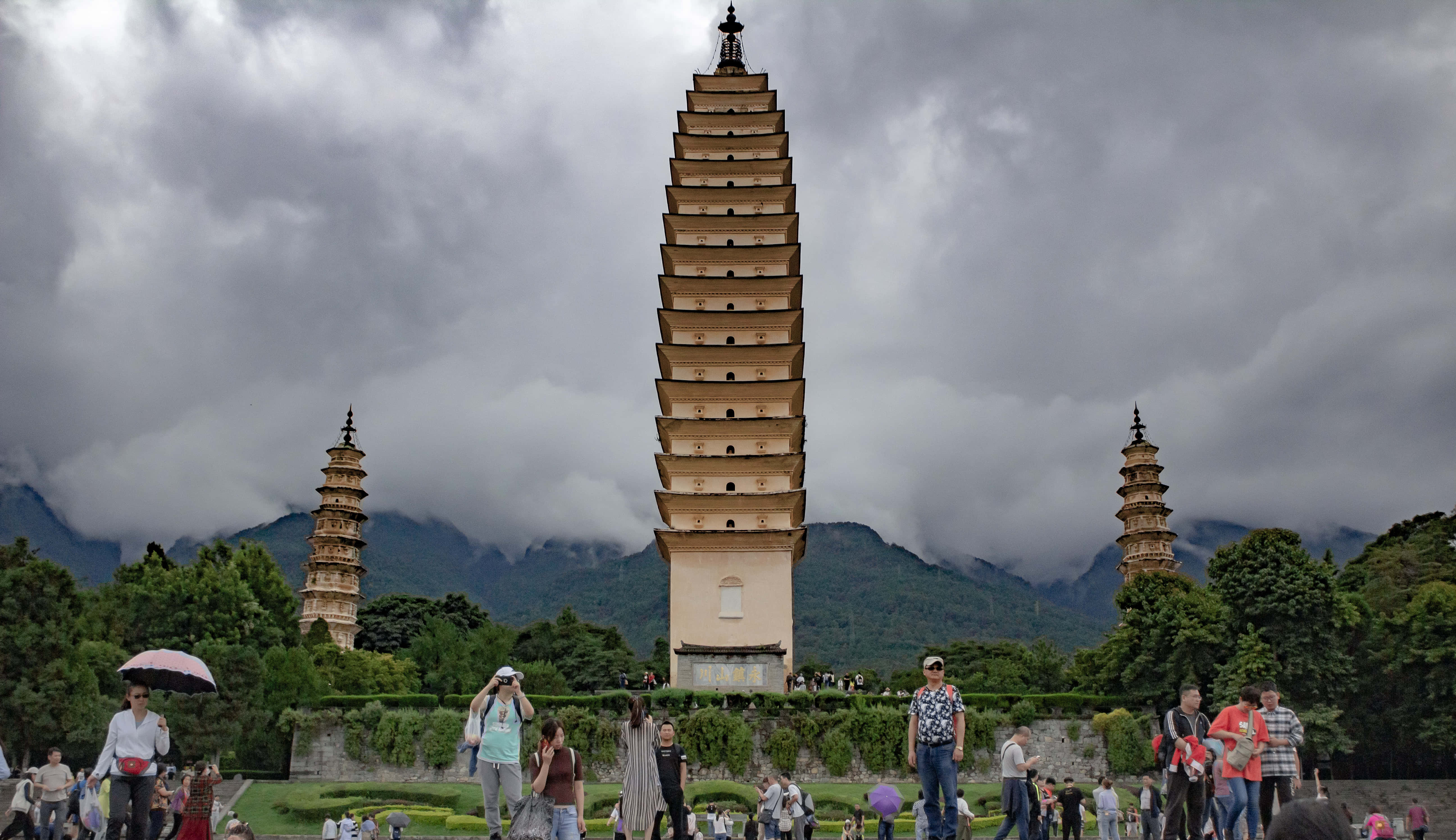 人物 桌布 壁紙 — 三座古塔在戲劇性的多雲天空下聳立，背景是霧氣繚繞的山脈，遊客正在探索場地。
