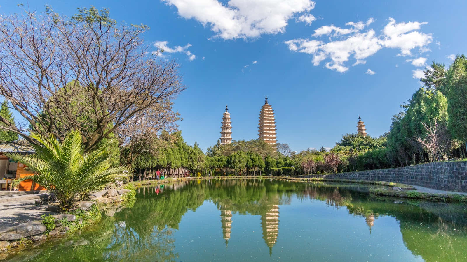 River View 桌布 壁紙 — 三座古塔在明亮的藍天下聳立，完美倒映在一個被茂密綠樹環繞的平靜池塘中。