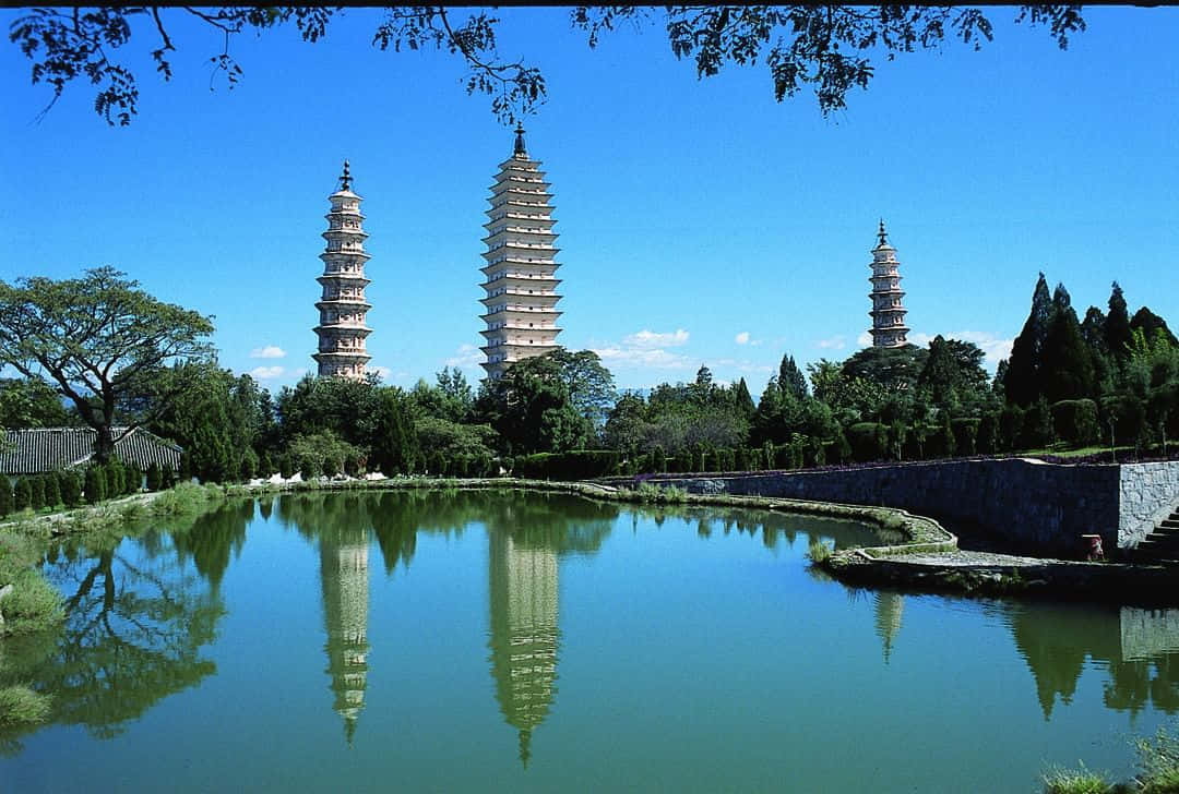 風景 桌布 壁紙 — 三座古塔在清澈的藍天下高聳，它們寧靜的倒影完美地映照在一個被茂密綠植環繞的平靜池塘中。