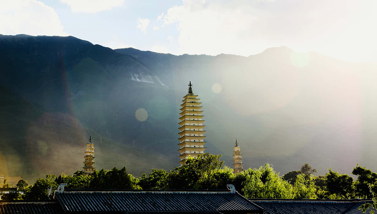 山脈 桌布 壁紙 — 三座古塔在茂密的綠樹之上聳立，背景是沐浴在金色日出光芒中的霧氣繚繞的山巒。