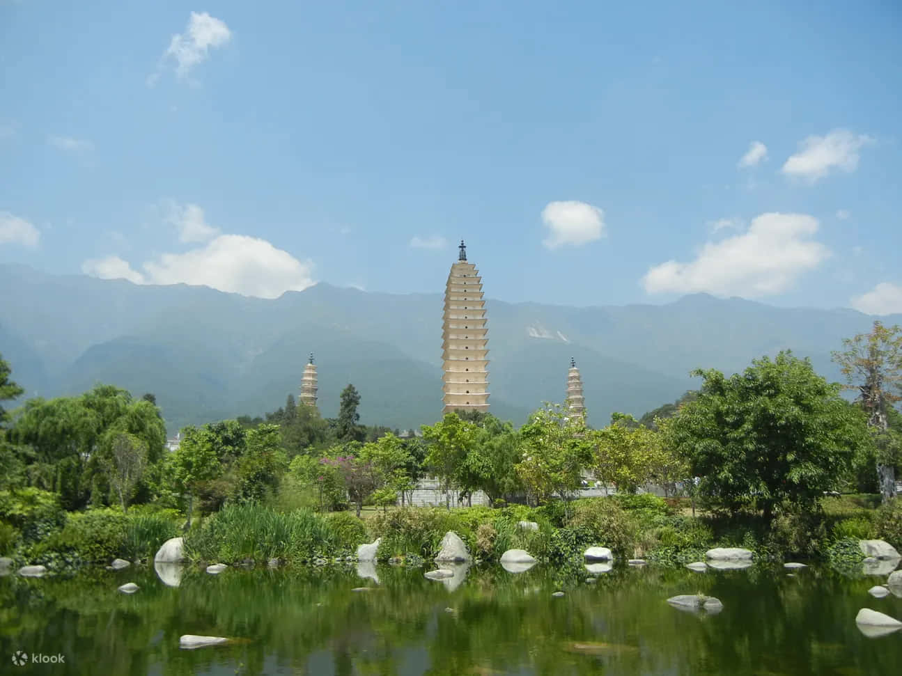風景 桌布 壁紙 — 三座古塔聳立在茂密的綠植和寧靜的池塘之上，池塘倒映著清澈的藍天和遠山。