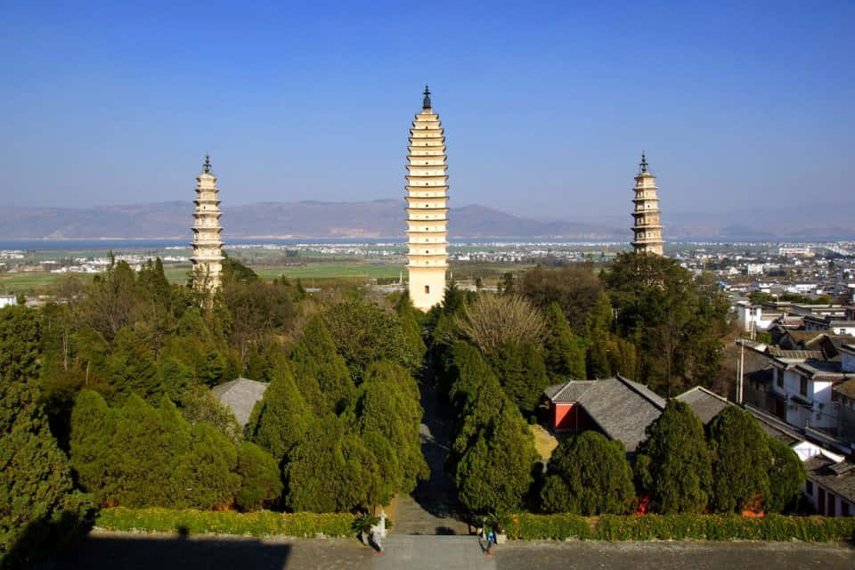 風景 桌布 壁紙 — 三座古塔聳立在茂密的綠樹之上，俯瞰著廣闊的城鎮和遠處的湖泊，天空湛藍清澈。
