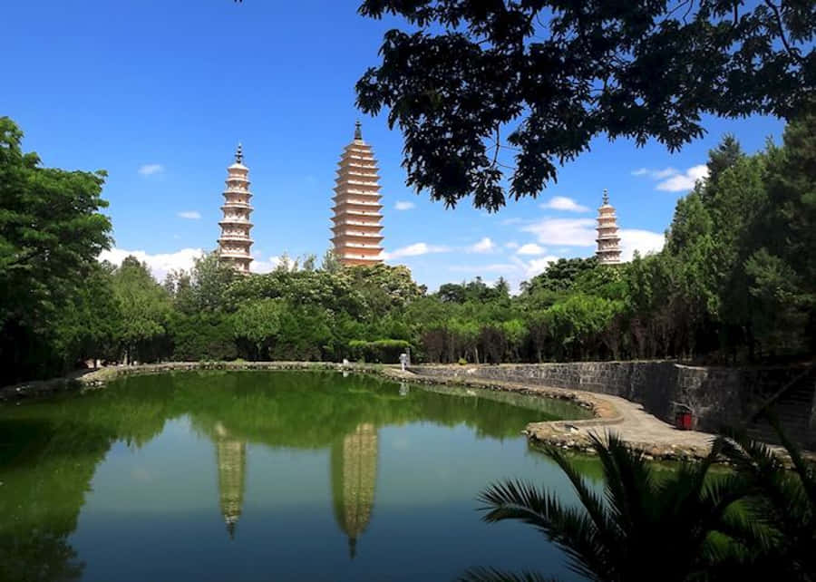 廟宇 桌布 壁紙 — 三座古塔聳立在茂密的綠樹之上，倒映在平靜的池塘中，上方是明亮的藍天。