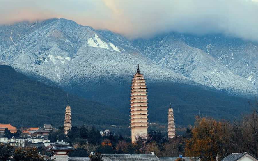 山脈 桌布 壁紙 — 三座古塔聳立於城鎮之上，背景是白雪覆蓋的山脈和陰雲密佈的天空。