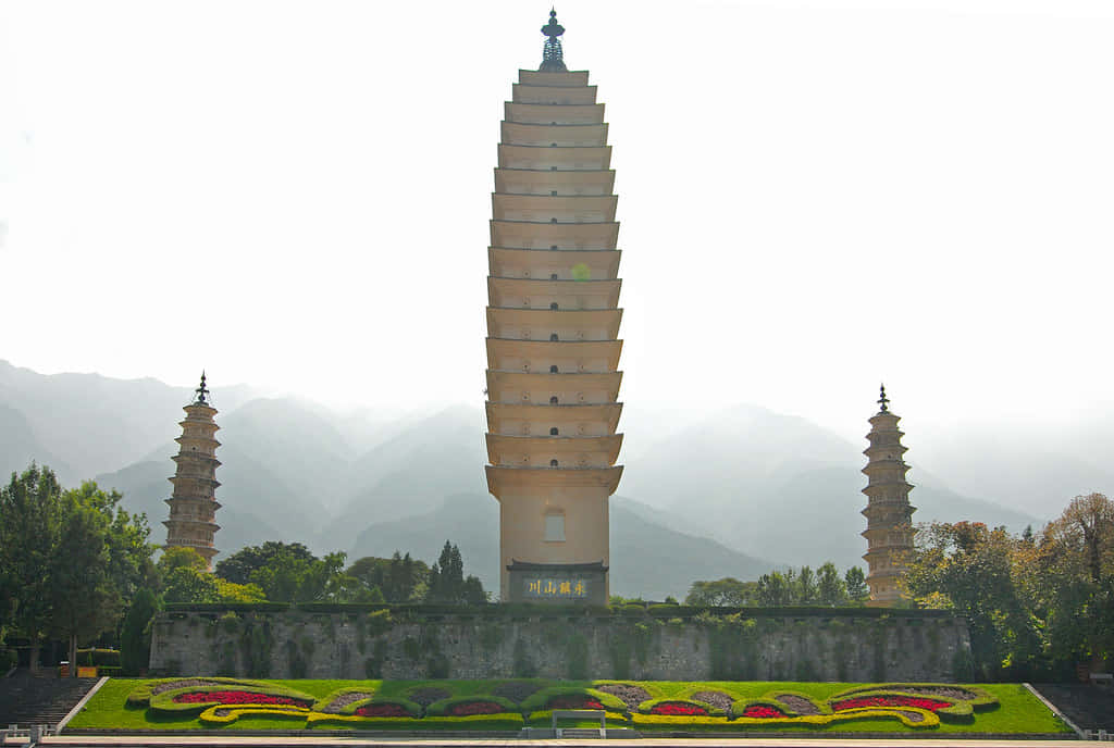 建築 桌布 壁紙 — 三座古老的多層寶塔在霧氣繚繞的山巒背景下雄偉地聳立，前景是色彩繽紛的花壇。