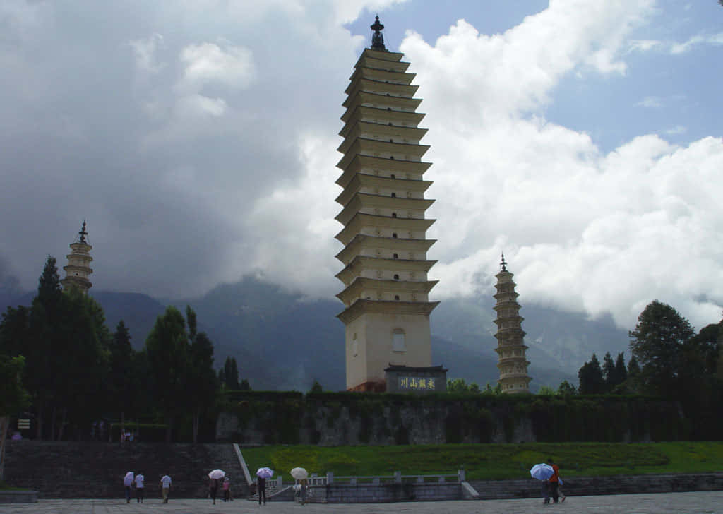 風景 桌布 壁紙 — 三座古老的層疊寶塔在陰雲密布的天空下聳立，背景是霧氣繚繞的山巒，下方有遊客撐著雨傘。