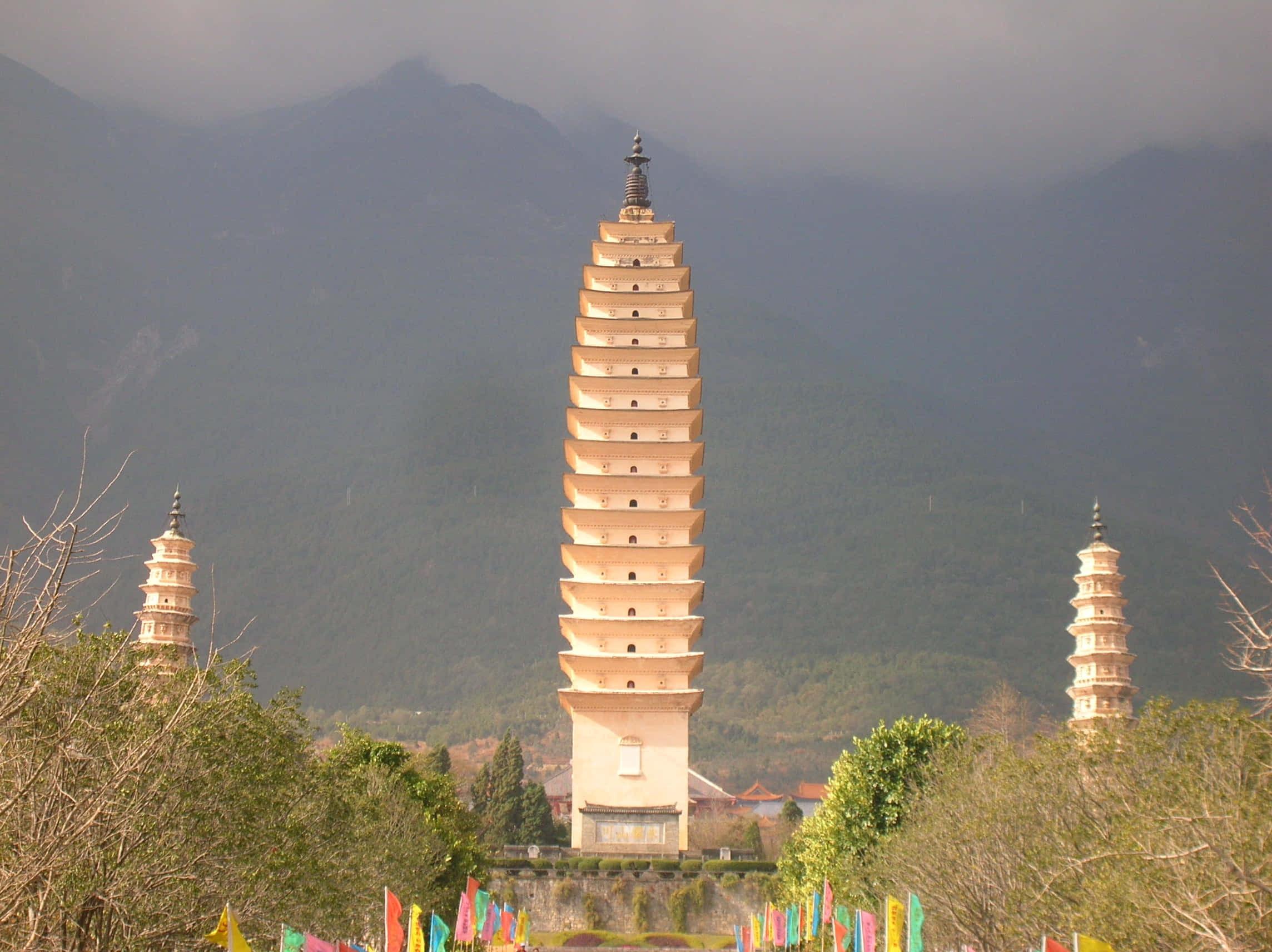 Buildings 桌布 壁紙 — 三座古老石塔在煙霧繚繞的山巒背景下巍然聳立，前景中五彩經幡隨風飄揚。
