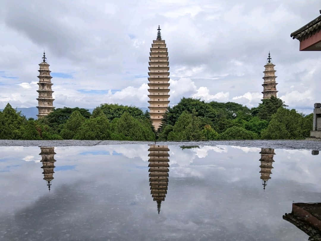 建築 桌布 壁紙 — 三座古老石塔矗立在茂密綠樹後方，完美倒映在平靜的池塘中，天空多雲。