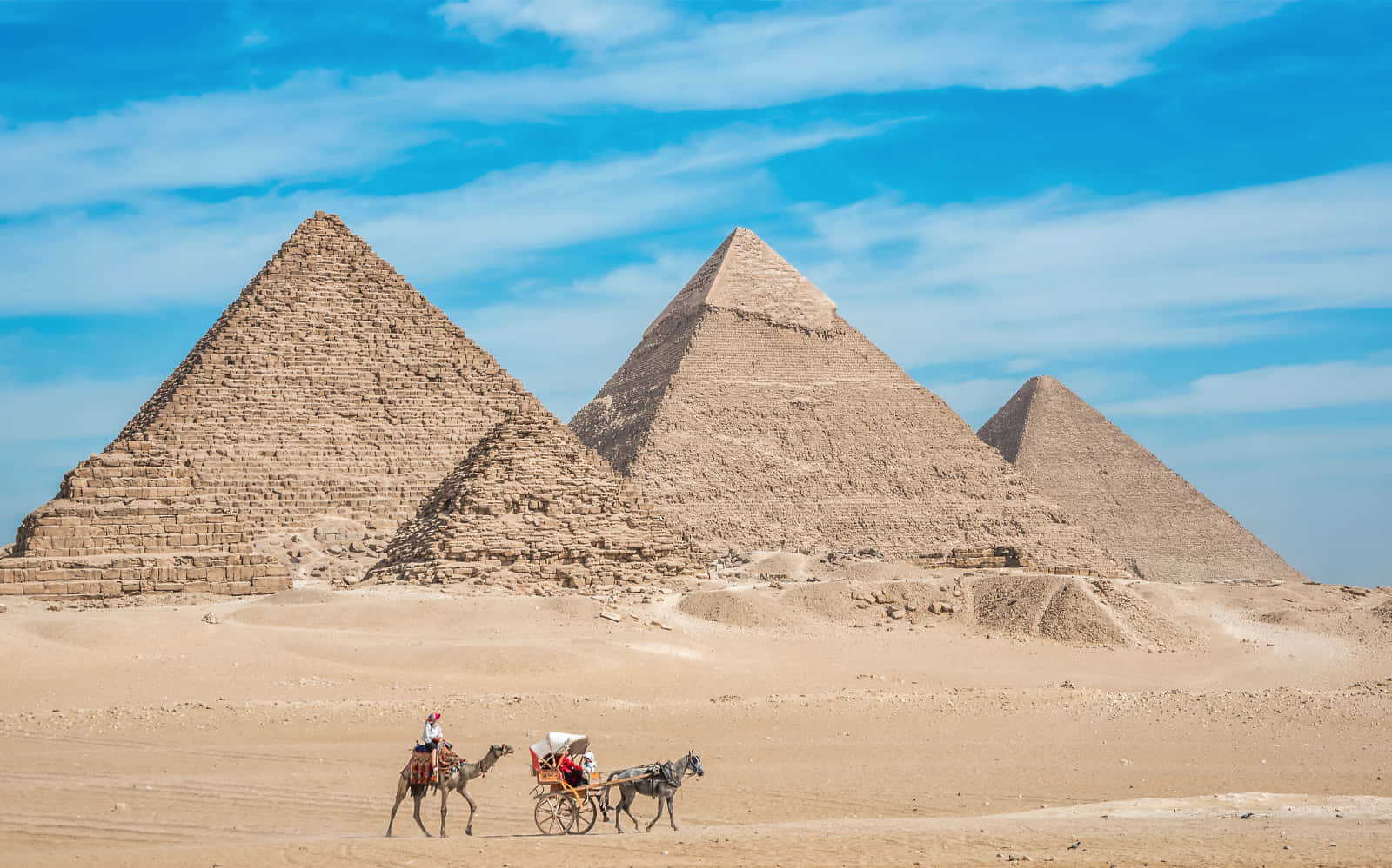 Camel 桌布 壁紙 — 三座巨大的金字塔從金色沙漠中升起，在明亮的藍天下有縷縷白雲，前景中一名騎駱駝的人和馬車穿行而過。