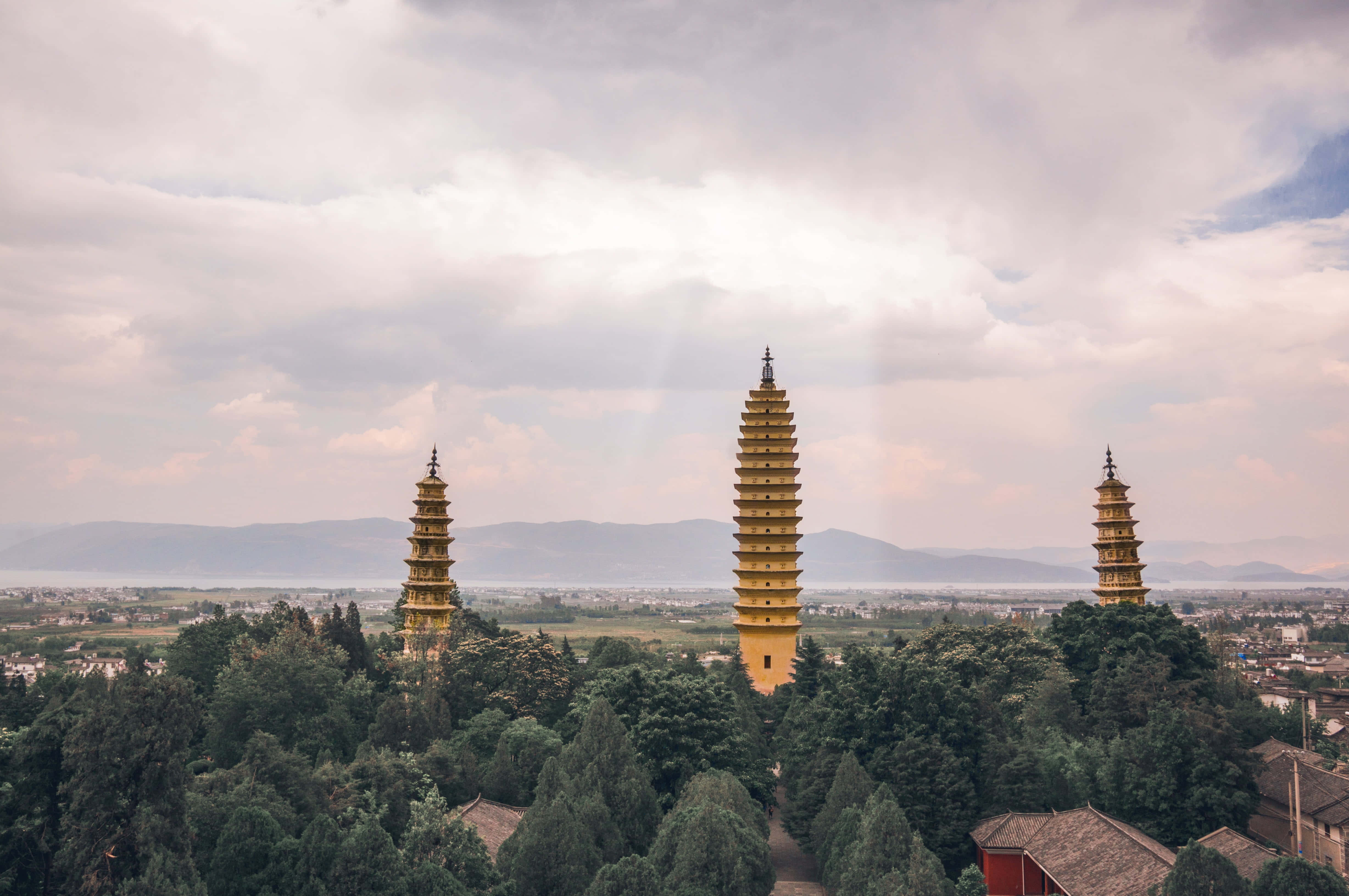 Cloudy Sky 桌布 壁紙 — 三座金色寶塔在陰天中聳立於茂密的綠樹之上，背景有遠山。