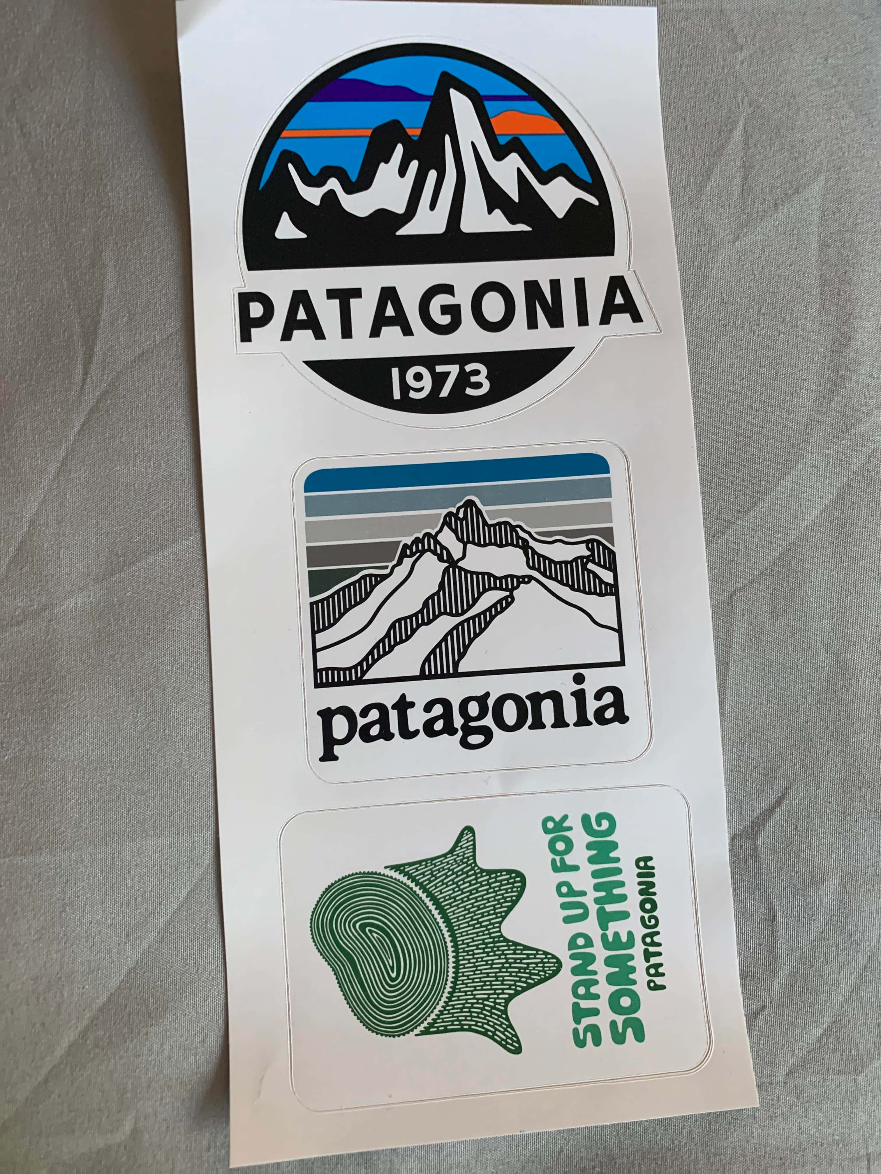 Patagonia 桌布 壁紙 — 三張Patagonia品牌貼紙，上面有山脈標誌和樹木年輪設計，放在白色紙張上，背景為灰色布料。