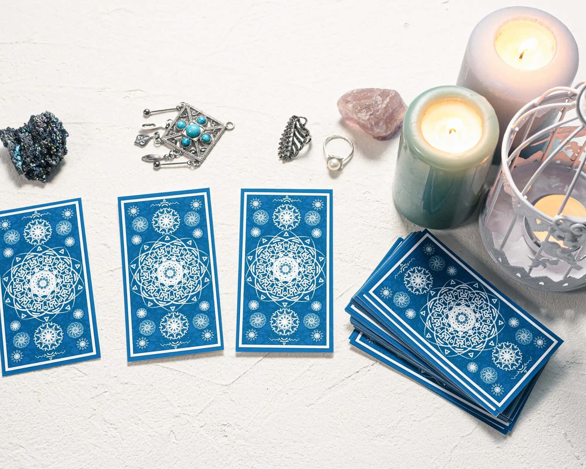 Tarot Card 桌布 壁紙 — 三張帶有白色曼陀羅圖案的藍色塔羅牌平放在一堆旁邊，周圍是水晶首飾和點燃的蠟燭，置於有紋理的白色表面上。