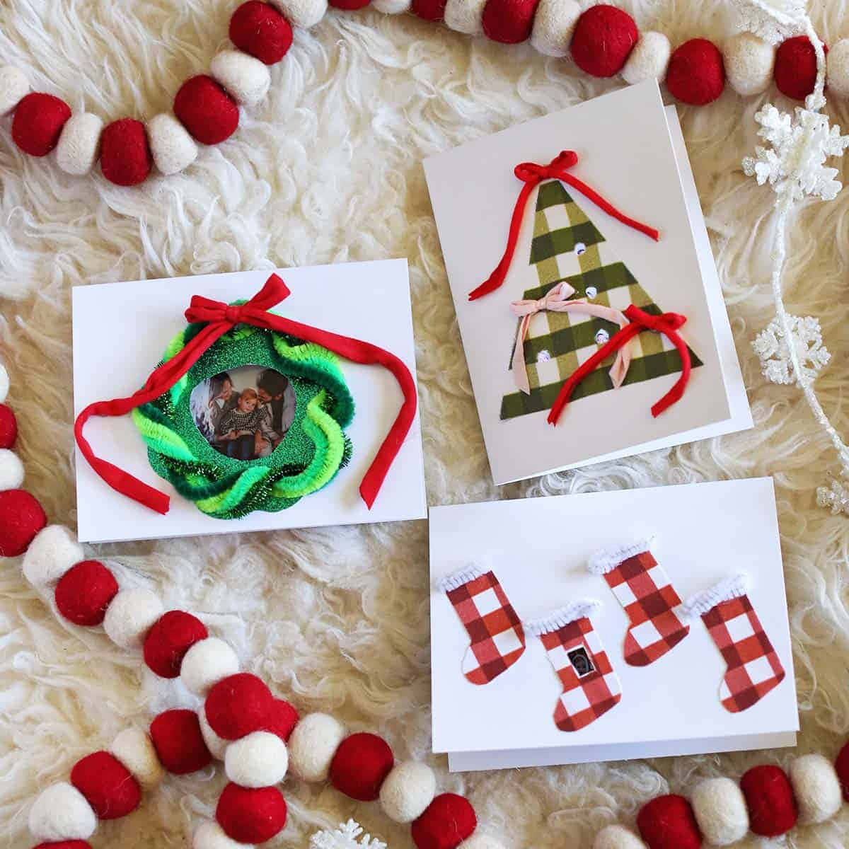 Christmas Card 桌布 壁紙 — 三張手工聖誕卡，設計有花環、聖誕樹和襪子，放在一張毛絨白色地毯上，周圍環繞著紅色和白色的絨球花環。
