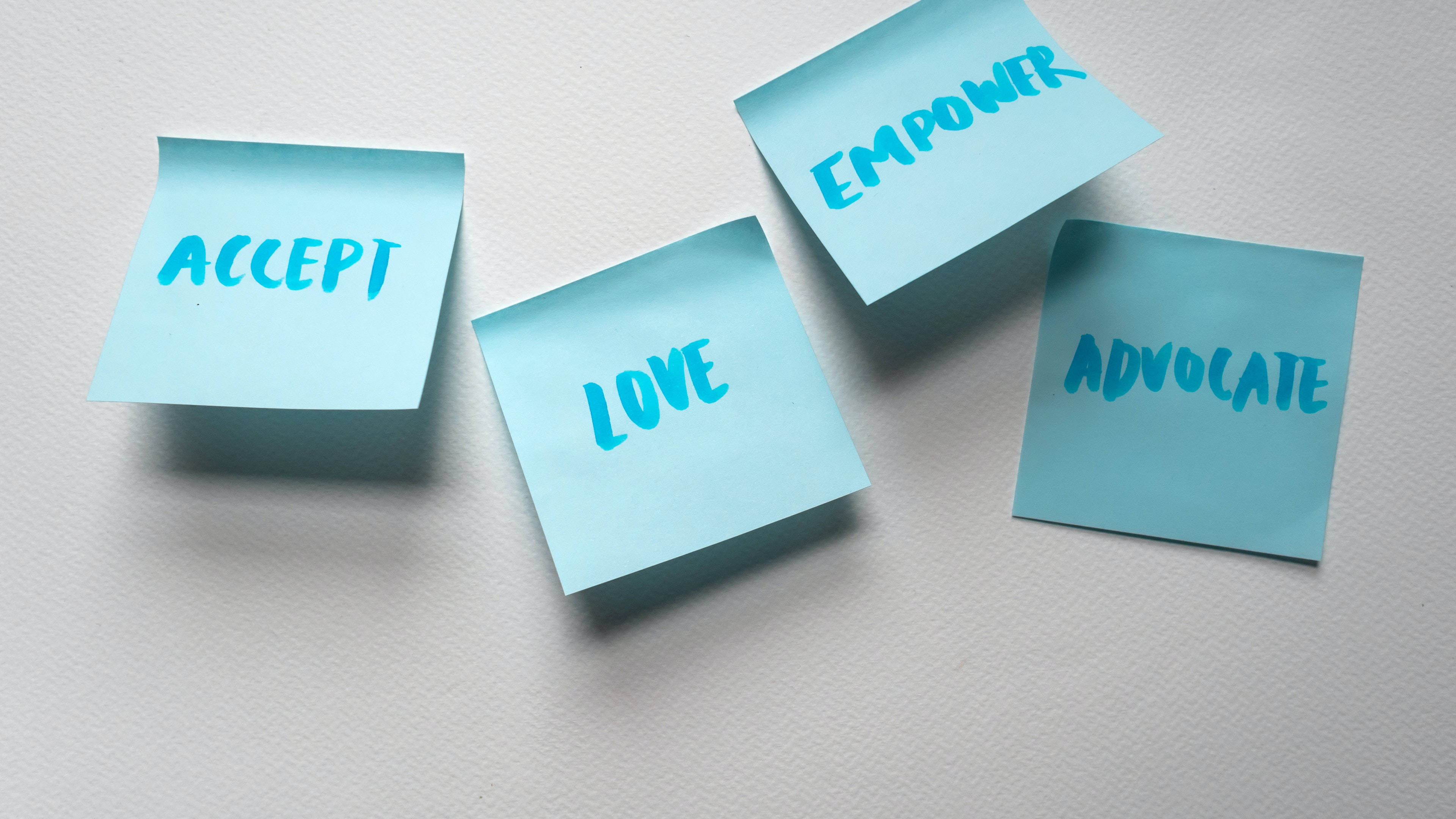 桌布 壁紙 — 三張淺藍色便利貼，上面有手寫文字 accept love empower advocate，排列在帶紋理的白色表面上。
