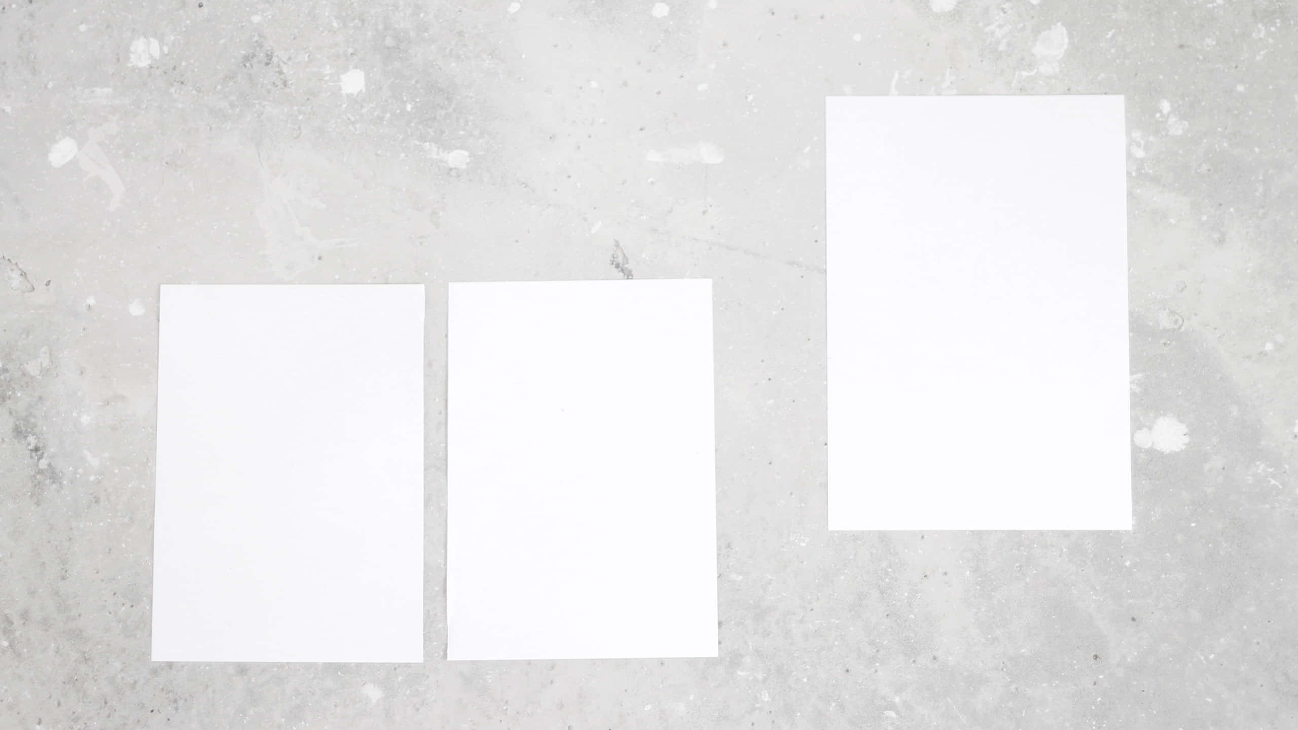 Soft White Aesthetic 桌布 壁紙 — 三張空白白紙放在帶有紋理的灰色混凝土表面上，散落著油漆濺點。
