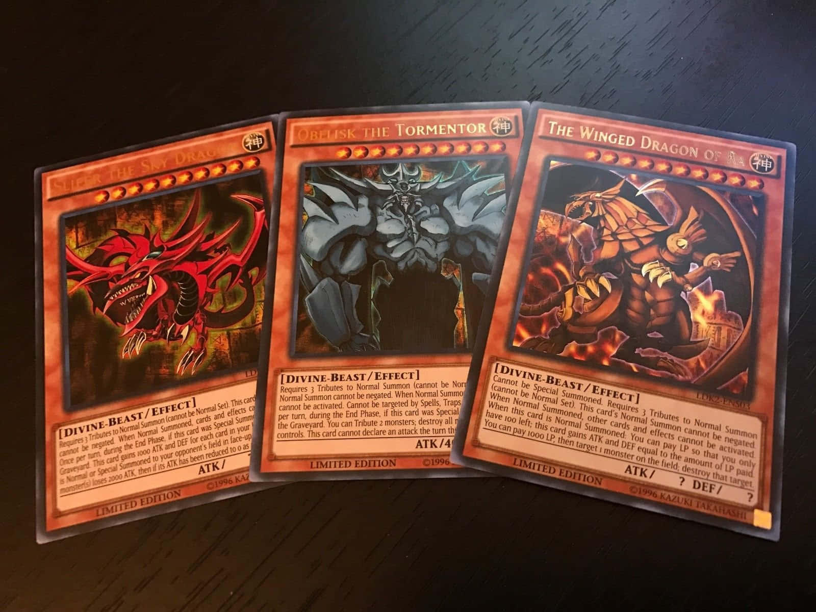 The Winged Dragon Of Ra 桌布 壁紙 — 三張遊戲王集換式卡牌描繪著火焰紅龍神獸，扇形散開在深色木桌上。