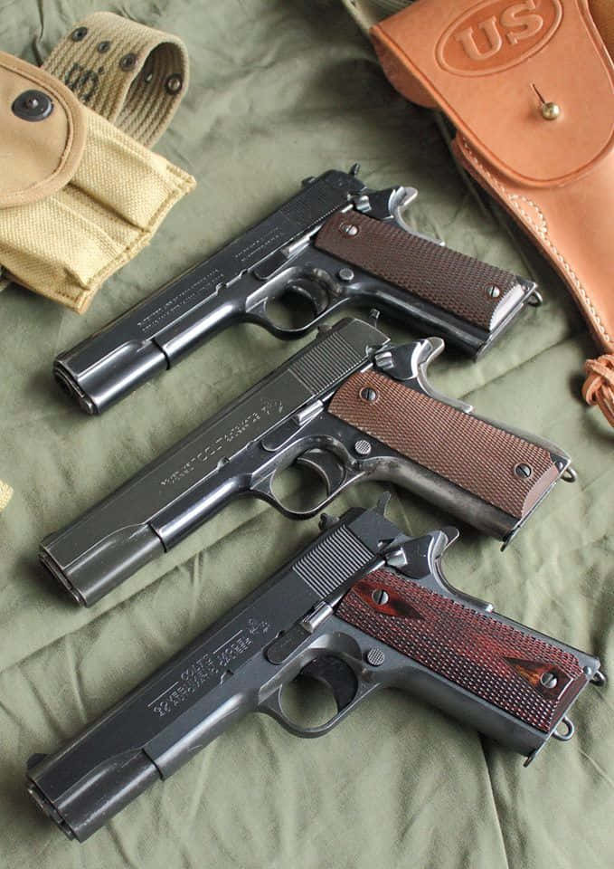 pistol 桌布 壁紙 — 三把軍用風格手槍，木製握把，放在橄欖色布料上，旁邊有棕褐色和棕色皮槍套。