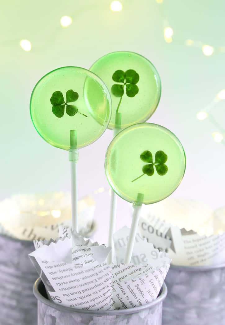Four Leaf Clover 桌布 壁紙 — 三支半透明綠色棒棒糖，內嵌四葉草，在模糊的仙女燈和報紙背景上柔和地發光。