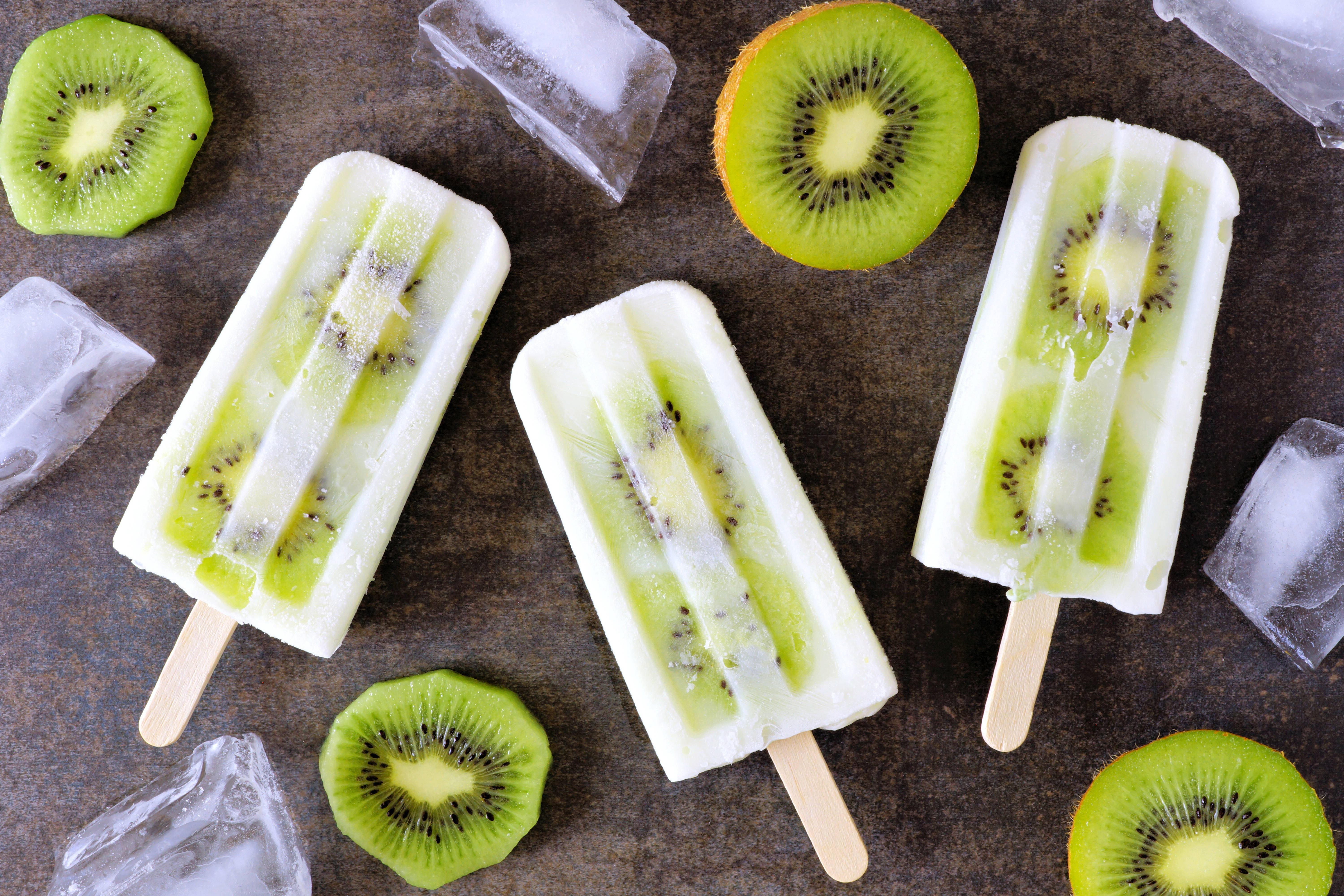 Fruit 桌布 壁紙 — 三支奇異果冰棒，可見水果切片，旁邊放著新鮮奇異果圓片和冰塊，置於深色紋理表面上。