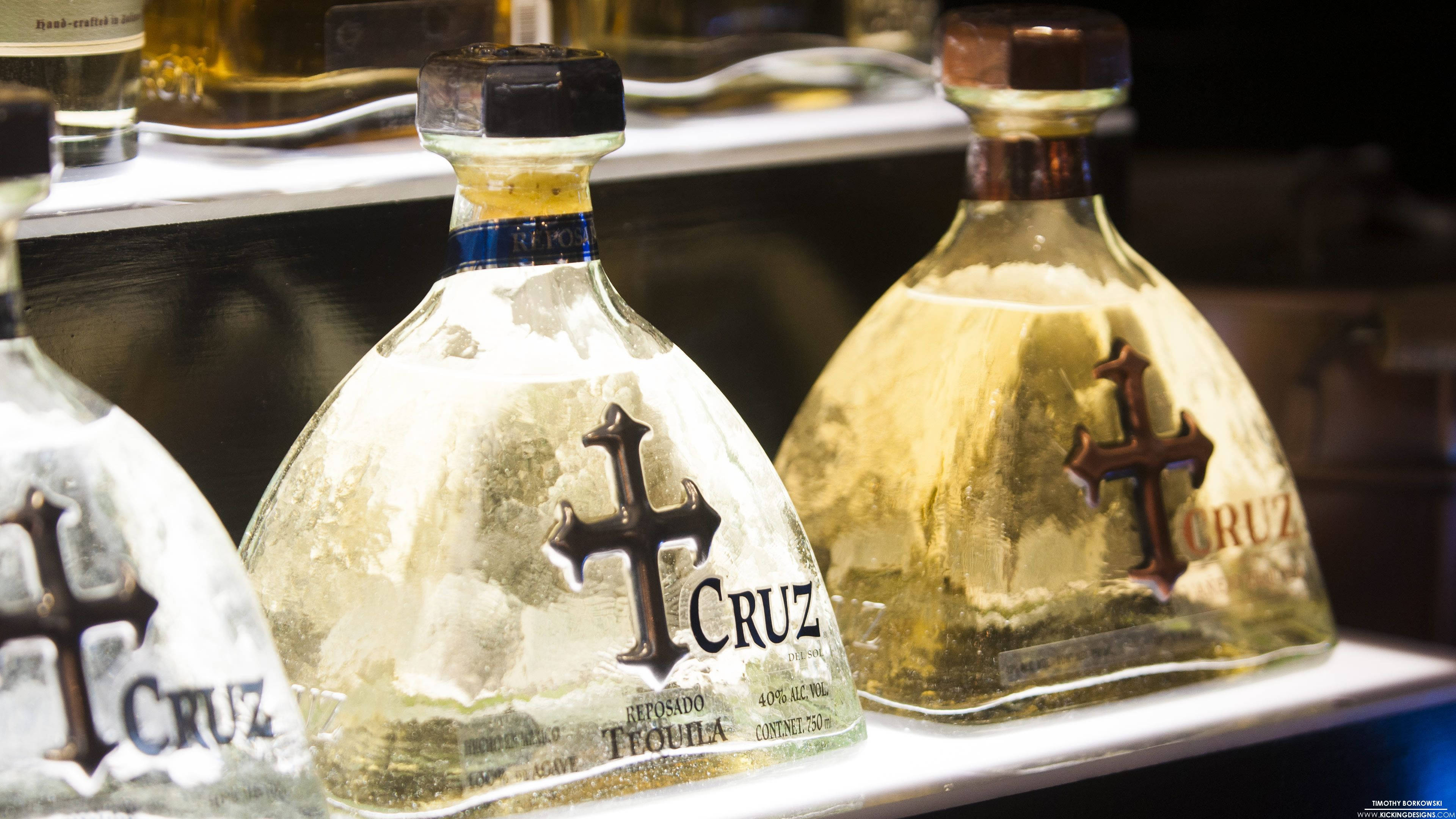 alcohol 桌布 壁紙 — 三支帶有十字標誌的Cruz龍舌蘭酒瓶，在溫暖燈光下放在酒吧架上，展示金色液體和精細標籤。