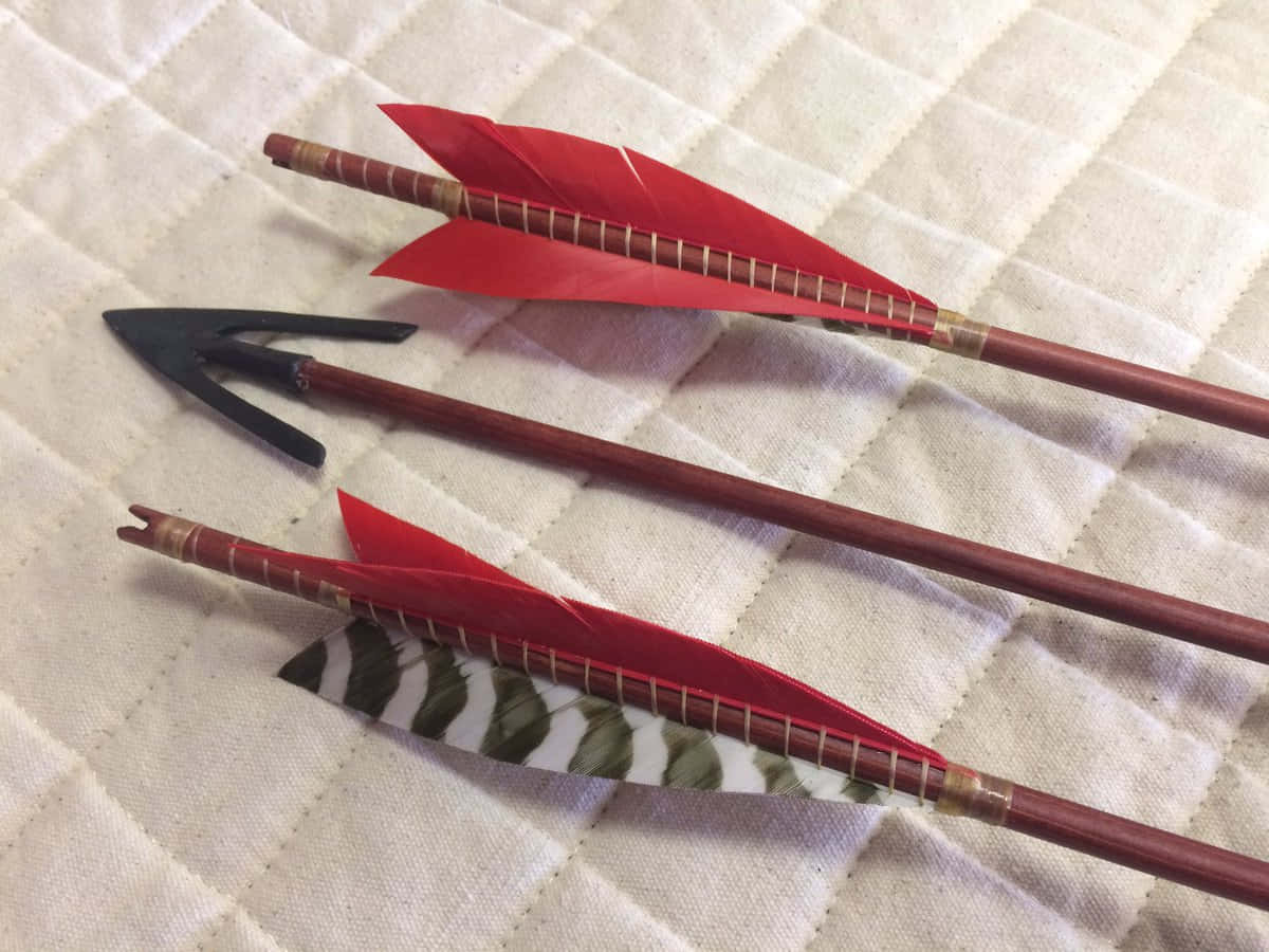 Arrow 桌布 壁紙 — 三支帶有紅色和圖案箭羽的箭斜放在奶油色絎縫布料表面上。