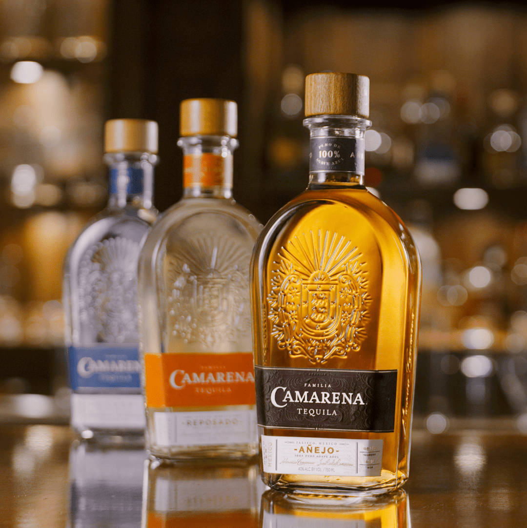 Tequila 桌布 壁紙 — 三支帶有金色瓶蓋和壓印標籤的Camarena龍舌蘭酒瓶，放置在反射的吧檯面上，背景是溫暖的散景效果。