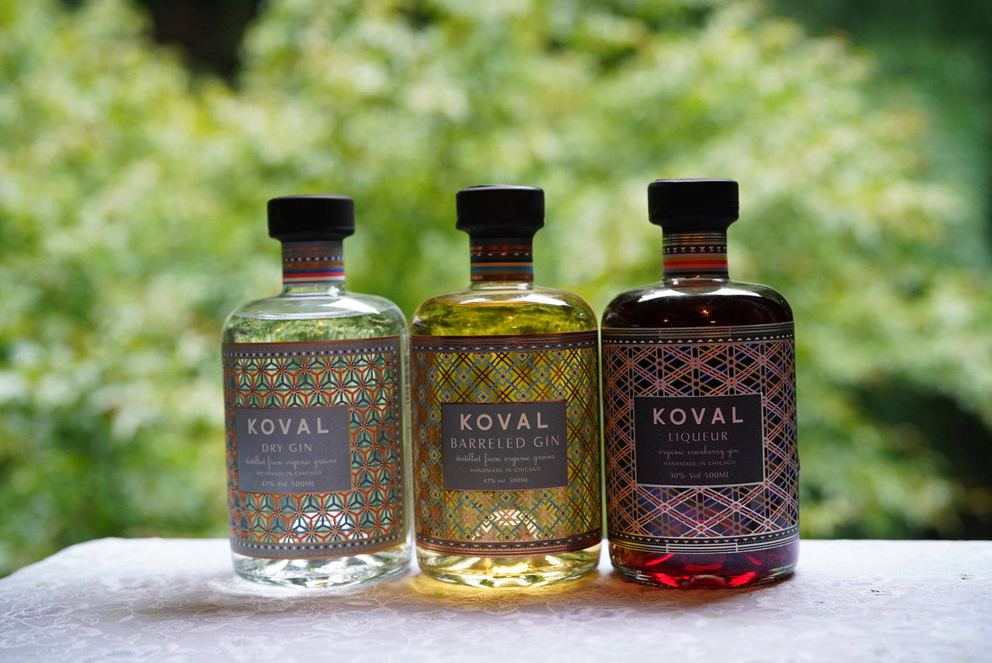 Whiskey 桌布 壁紙 — 三支裝飾標籤的Koval烈酒瓶，呈現清澈琥珀色和紅色色調，放在白色表面上，背景是柔和的綠色樹葉。