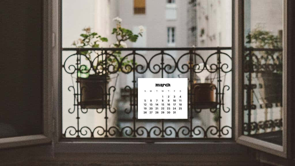March Desktop 桌布 壁紙 — 三月日曆掛在裝飾性的鍛鐵陽台欄杆上，旁邊有盆栽植物，背景是模糊的城市建築。