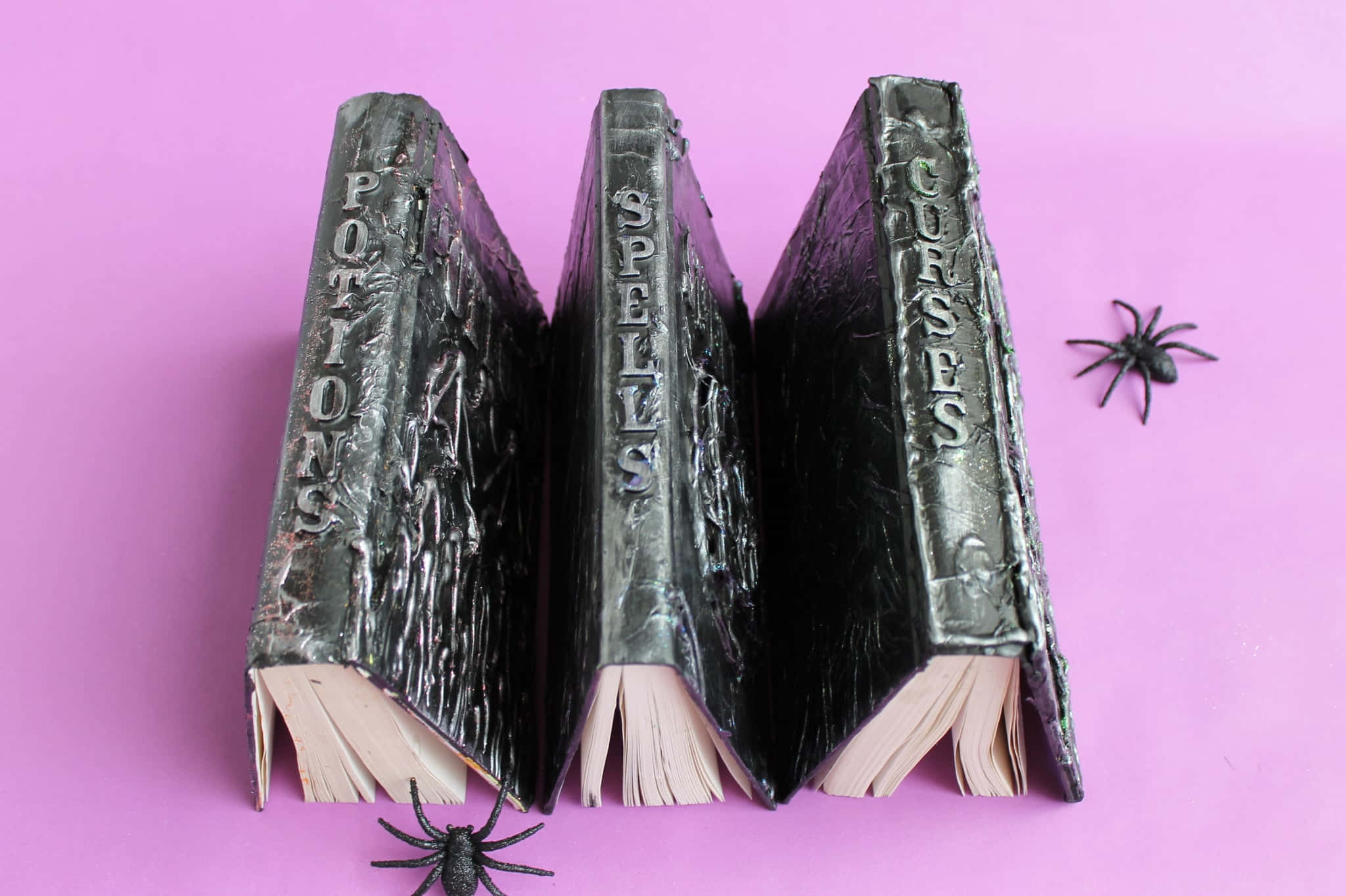 Halloween Books 桌布 壁紙 — 三本破舊的黑色咒語書，書名分別為《藥水》、《咒語》和《詛咒》，直立於鮮粉紅色表面上，附近有兩隻塑膠蜘蛛。