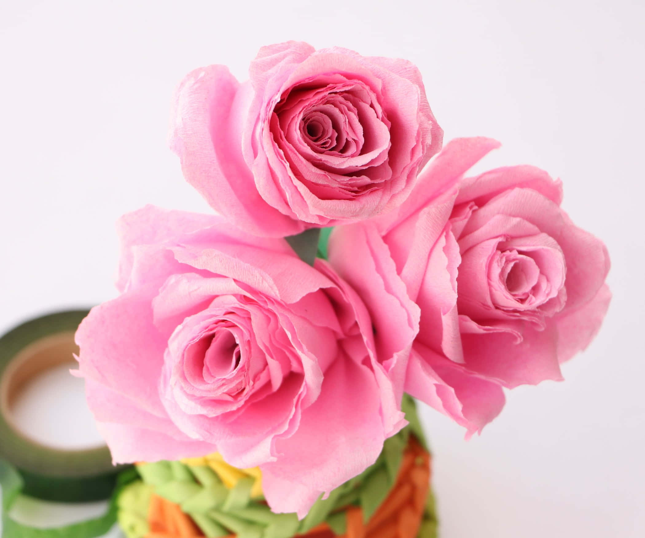 Rose Flower 桌布 壁紙 — 三朵精緻的粉紅色紙玫瑰在色彩繽紛的編織花瓶中盛開，旁邊有一卷綠色膠帶，背景是潔淨的白色。