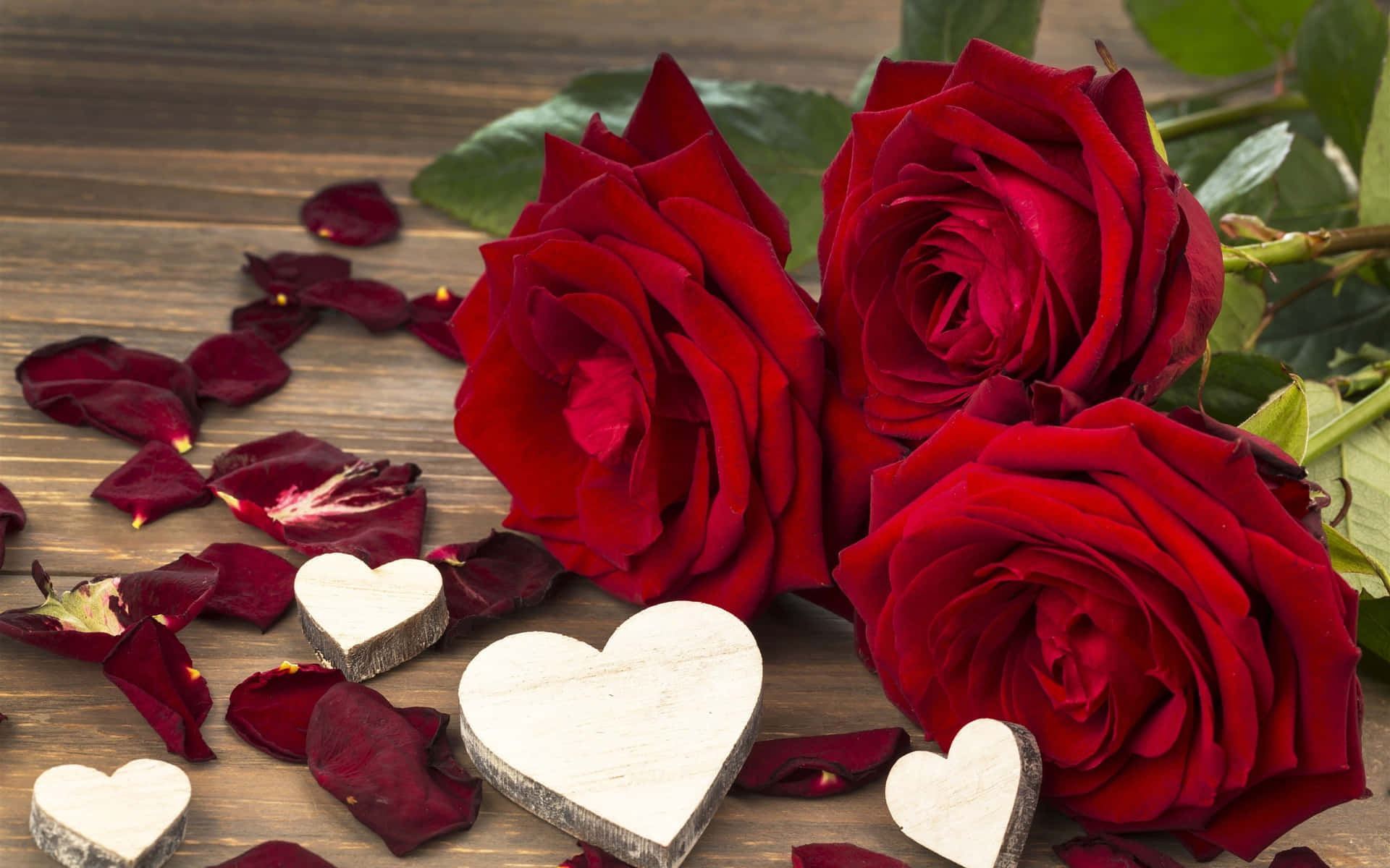 Roses 桌布 壁紙 — 三朵鮮豔的紅玫瑰，散落的花瓣和木製心形，放置在質樸的木質表面上。