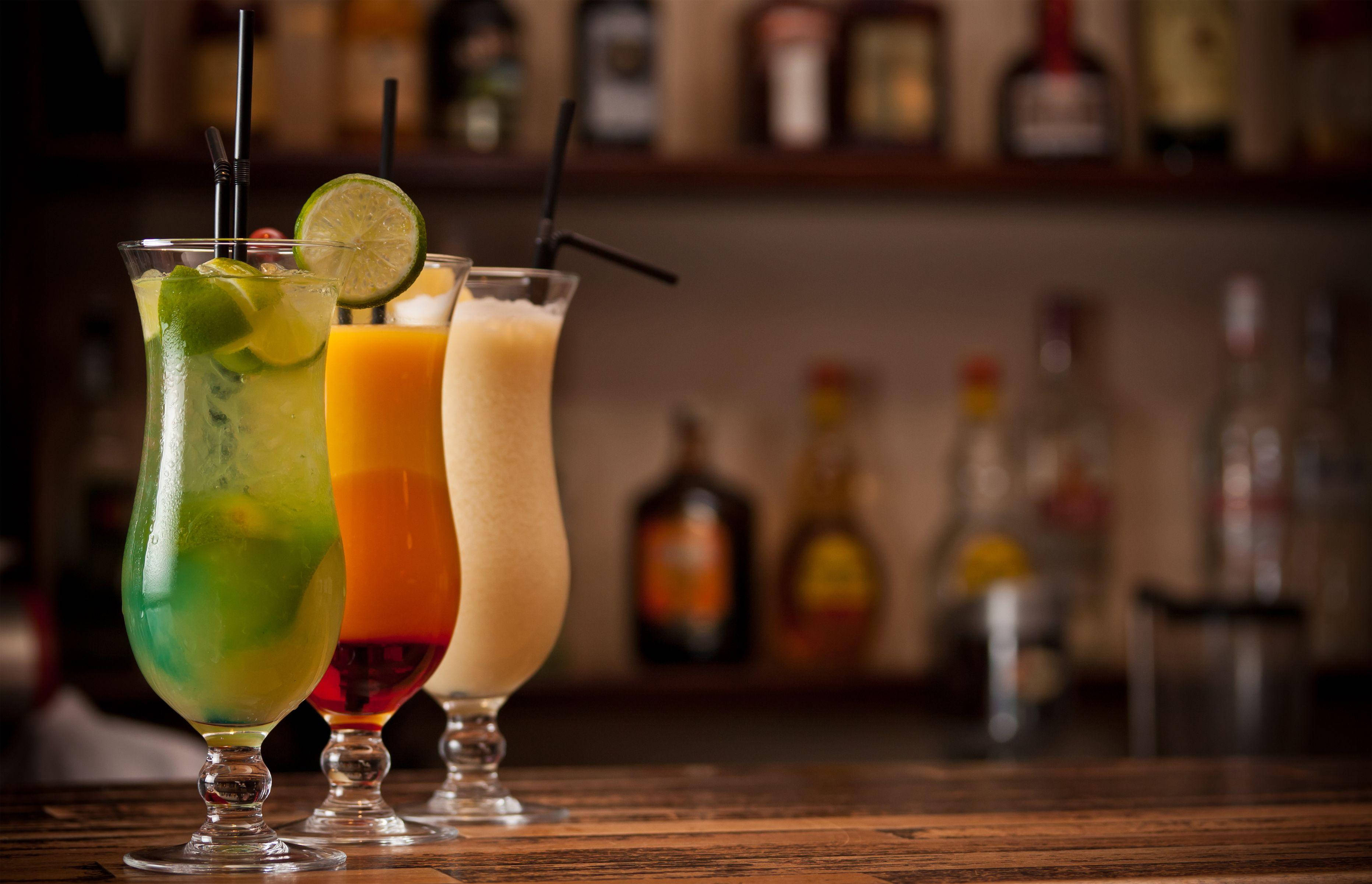 Bar 桌布 壁紙 — 三杯充滿活力的熱帶雞尾酒，配上青檸裝飾，放在木製吧檯上，背景有模糊的酒瓶。
