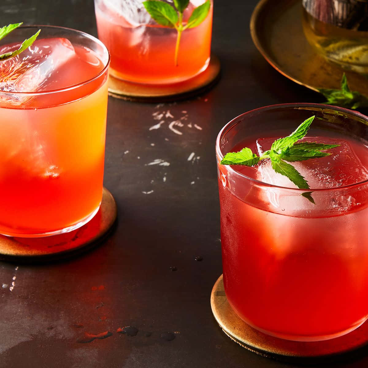 alcohol 桌布 壁紙 — 三杯充滿活力的紅色雞尾酒，搭配薄荷和冰塊，放在木製杯墊上，背景是深色紋理表面。