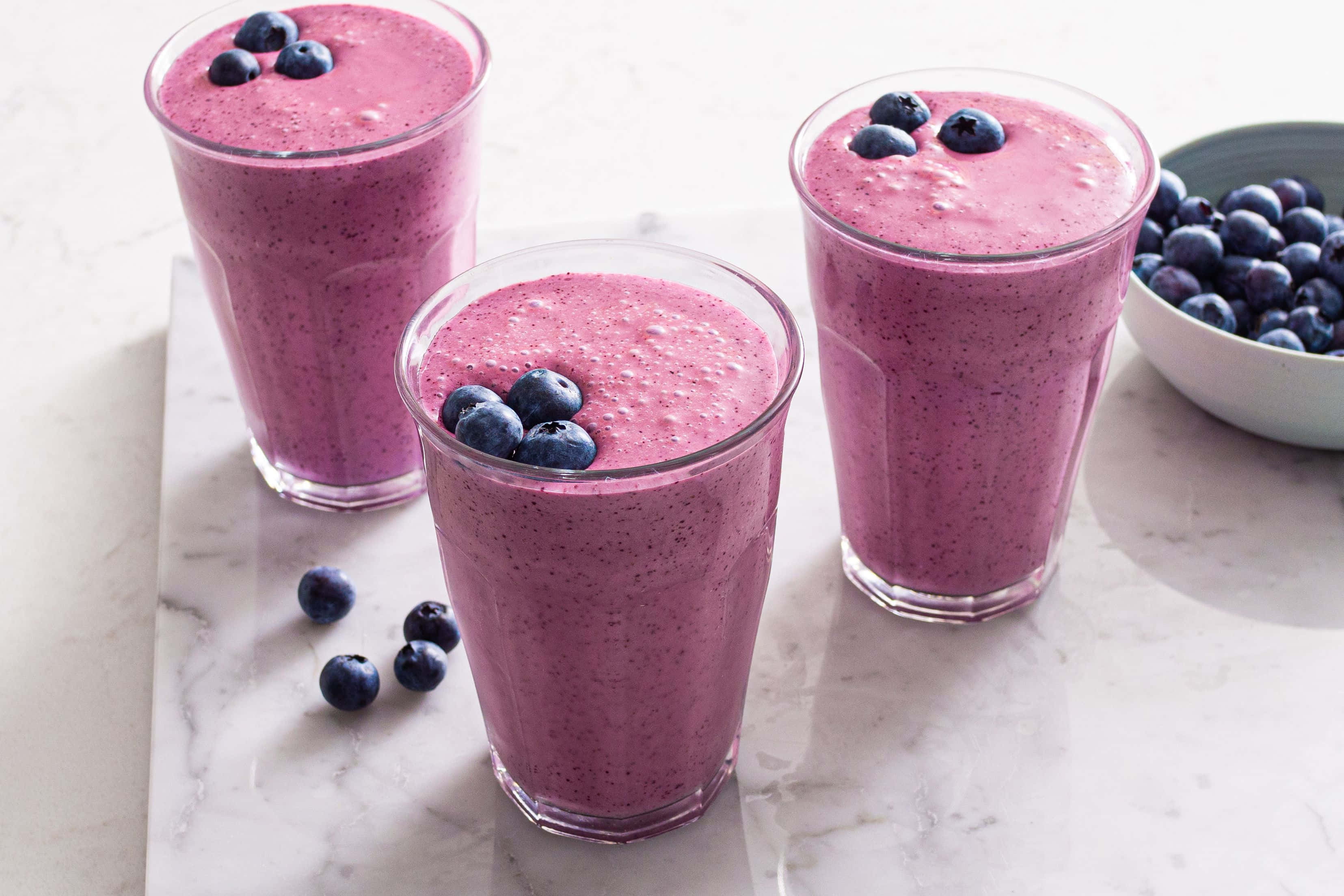 Blueberry Smoothie 桌布 壁紙 — 三杯充滿活力的紫色藍莓冰沙，上面放上新鮮藍莓，放在白色大理石表面上，旁邊有一碗莓果。
