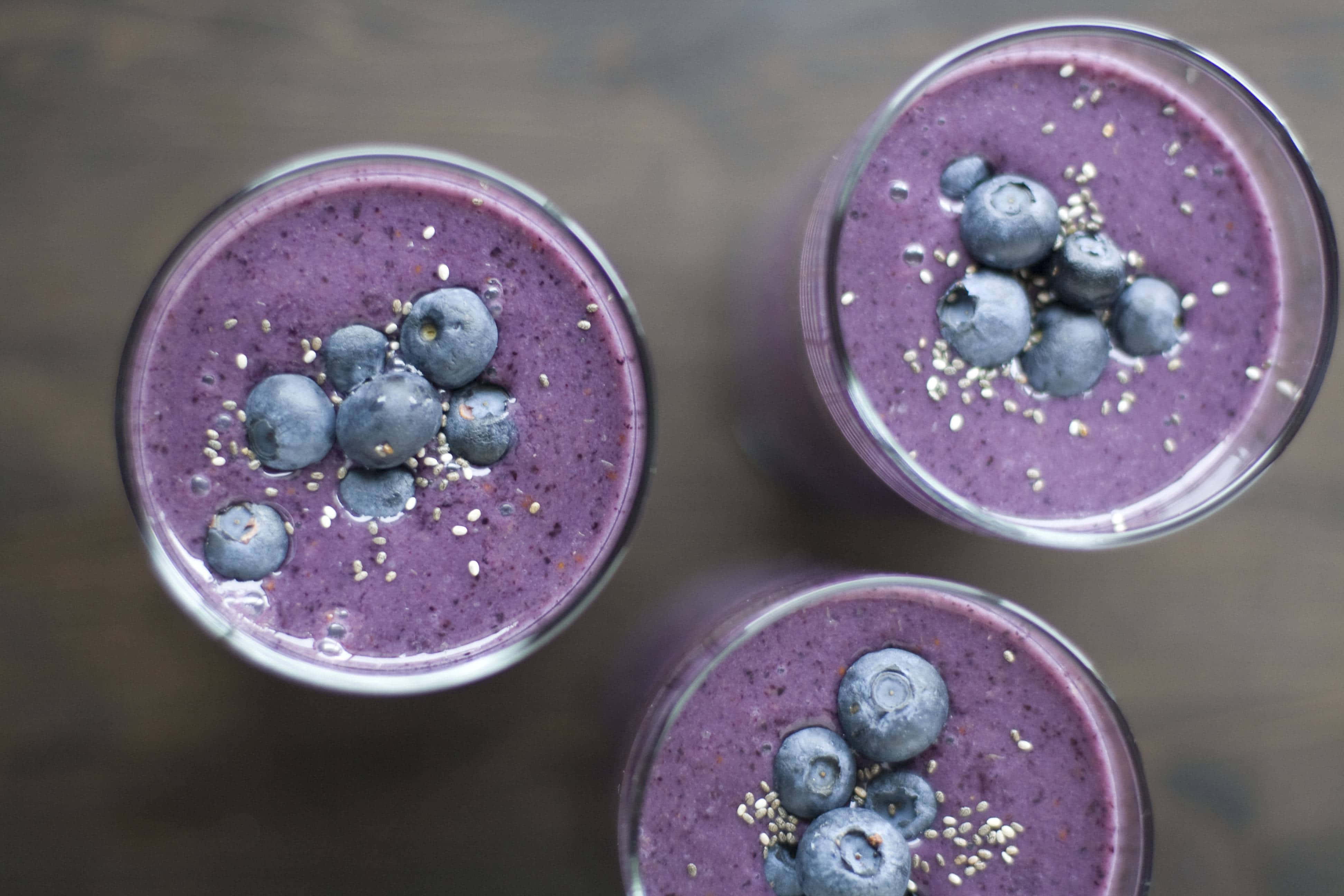 Blueberry Smoothie 桌布 壁紙 — 三杯充滿活力的紫色藍莓奶昔，上面放上新鮮藍莓和奇亞籽，置於質樸的木質表面上。