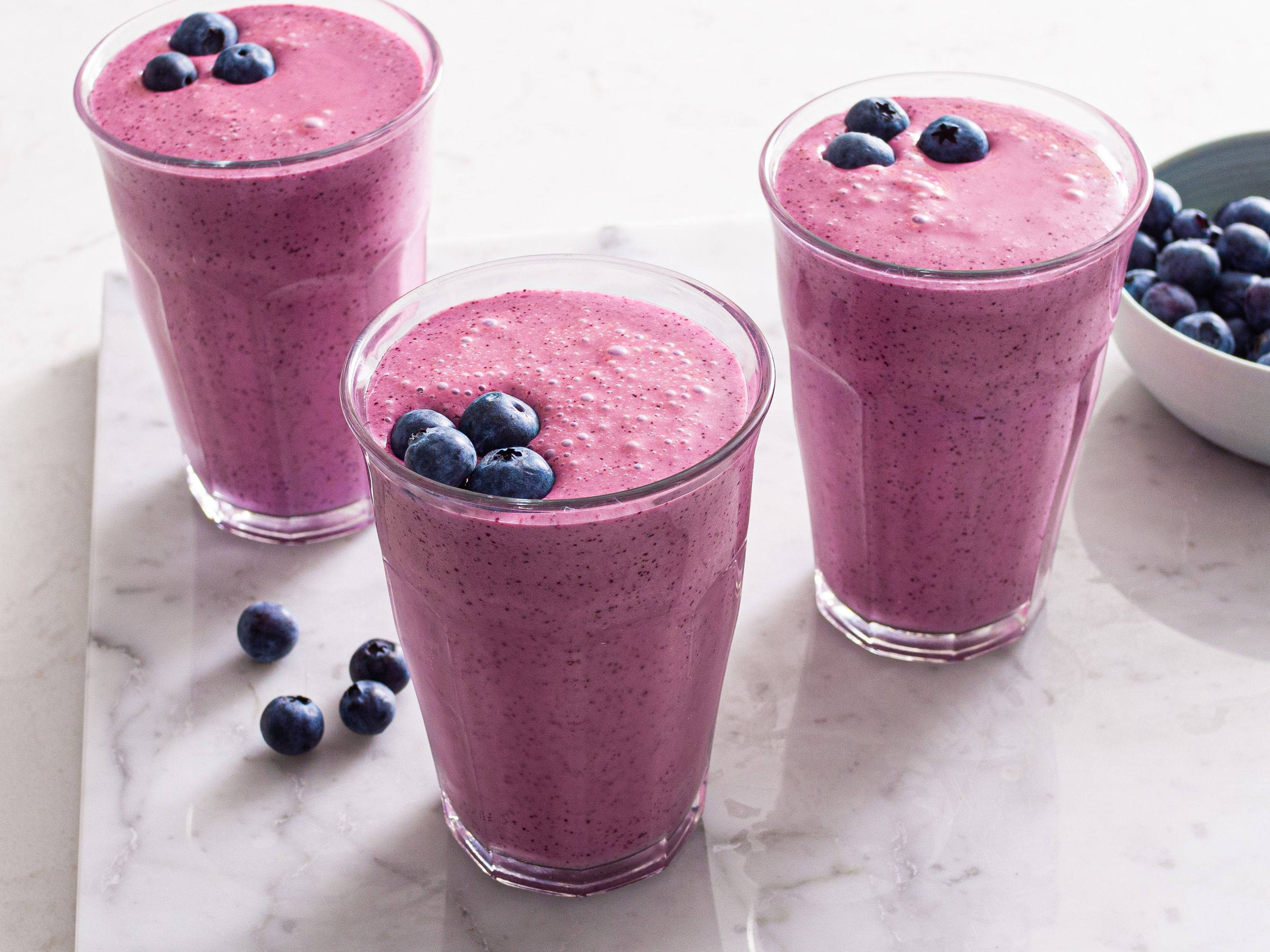Smoothie 桌布 壁紙 — 三杯充滿活力的紫藍色藍莓奶昔，放在透明玻璃杯中，上面放上新鮮藍莓，置於白色大理石表面上。
