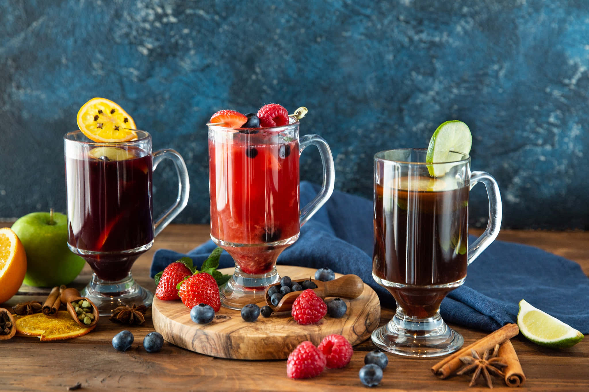 Food And Beverage 桌布 壁紙 — 三杯冒著熱氣的熱紅酒，飾以水果和香料，放在質樸的木板上，背景為帶紋理的藍色。