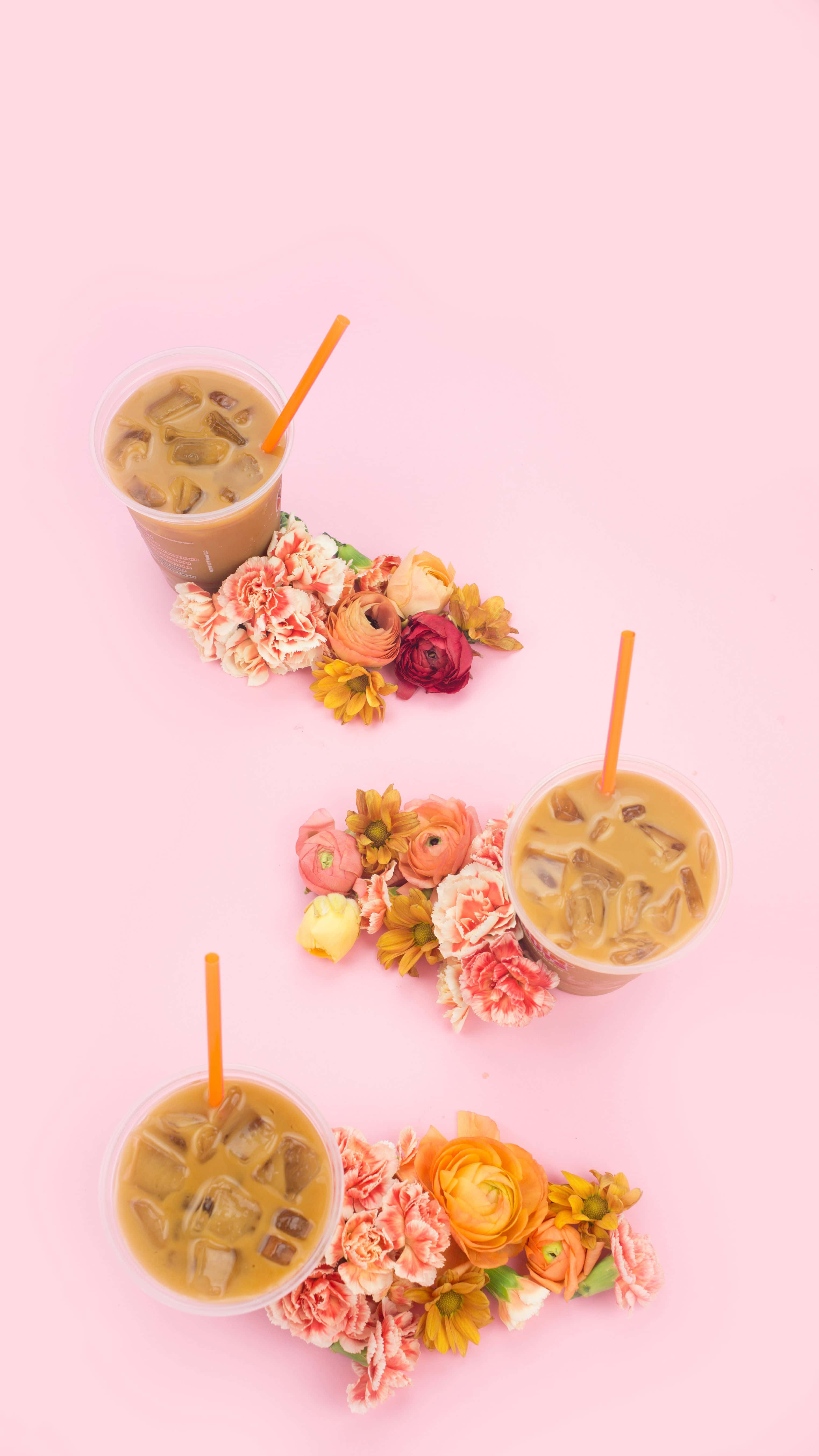 Dunkin Donuts 桌布 壁紙 — 三杯冰咖啡飲品，配上橙色吸管和花卉擺設，置於柔和粉紅色背景上。