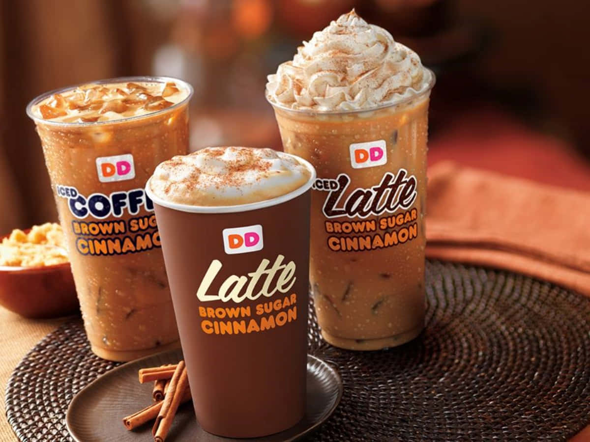 Dunkin Donuts 桌布 壁紙 — 三杯冰紅糖肉桂咖啡飲品，上面加上鮮奶油和肉桂，放在一個編織托盤上，旁邊有肉桂棒和一小碗顆粒。