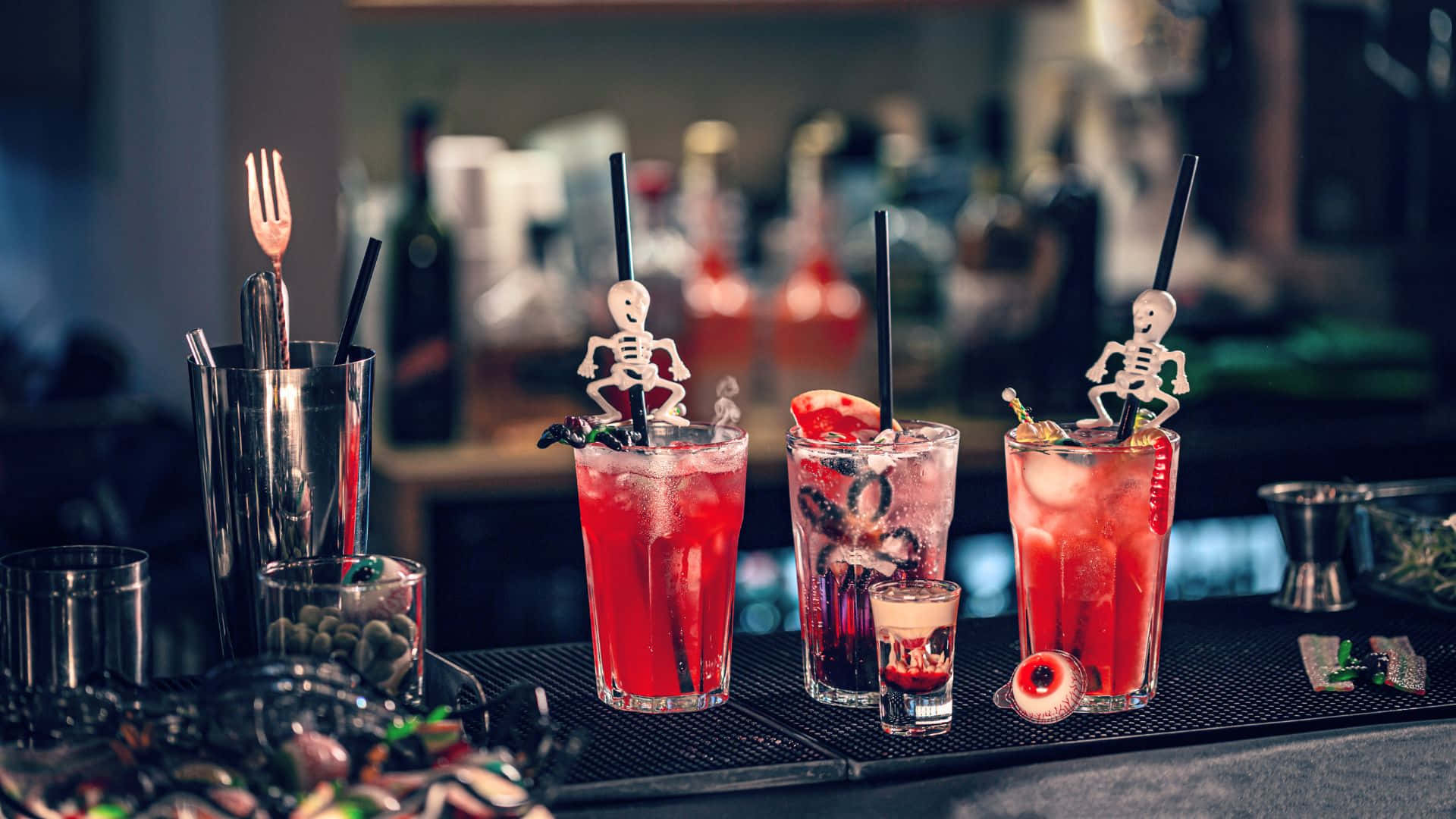 Spooky Cocktails 桌布 壁紙 — 三杯帶有骷髏裝飾棒和眼球裝飾的恐怖紅色雞尾酒，放在燈光昏暗的吧檯上。