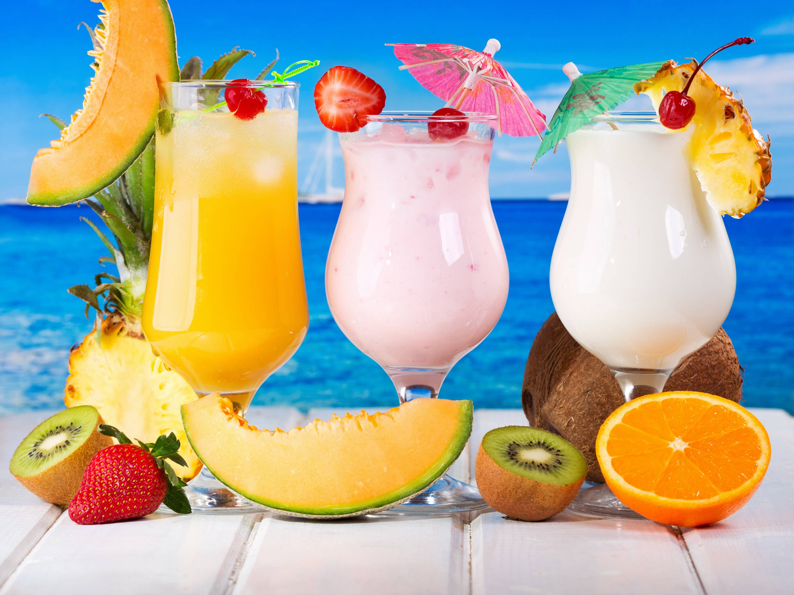 Tropical Drink 桌布 壁紙 — 三杯熱帶水果冰沙，飾以小傘和新鮮水果，放在白色木桌上，背景是明亮的藍色海洋。